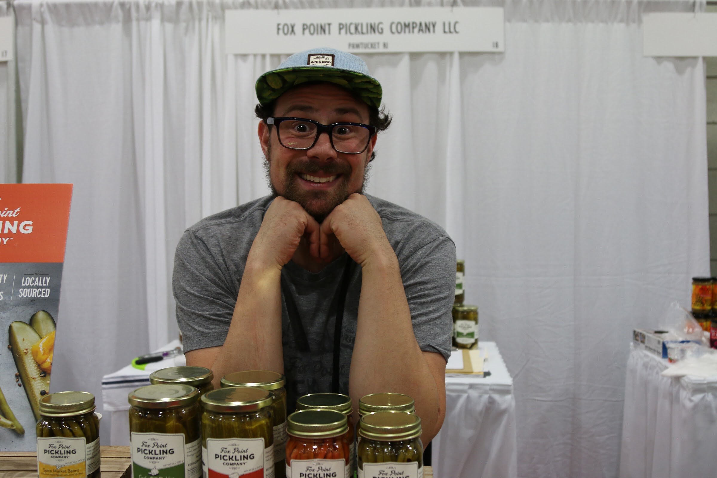 A grinning bearded man in glasses and a cap leaning on a counter lined with jars of Fox Point pickles.