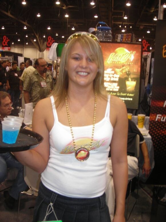 A smiling woman with sunglasses on her head wears a Banzai tank top and bead necklace, holding a tray of blue drinks in front of a lit Banzai sign.