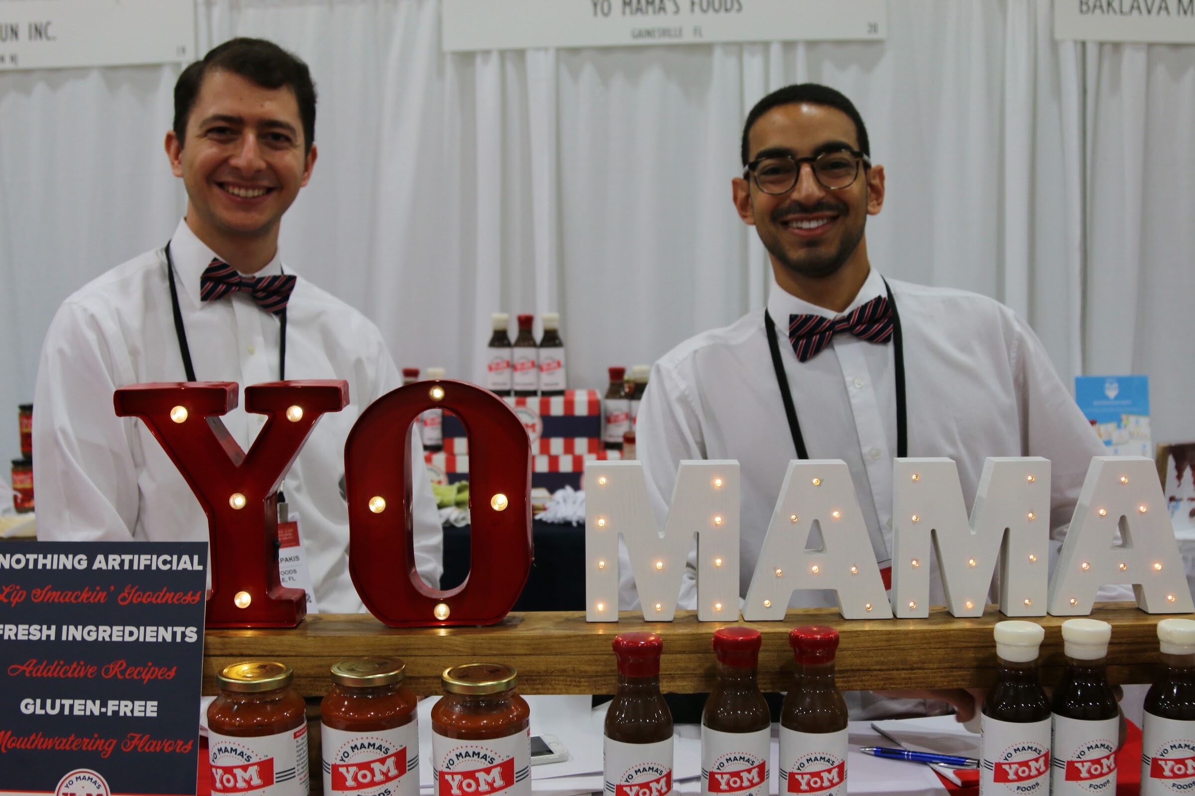 Two smiling men in bow ties behind a marquee-letter YO MAMA sign and rows of sauce jars.