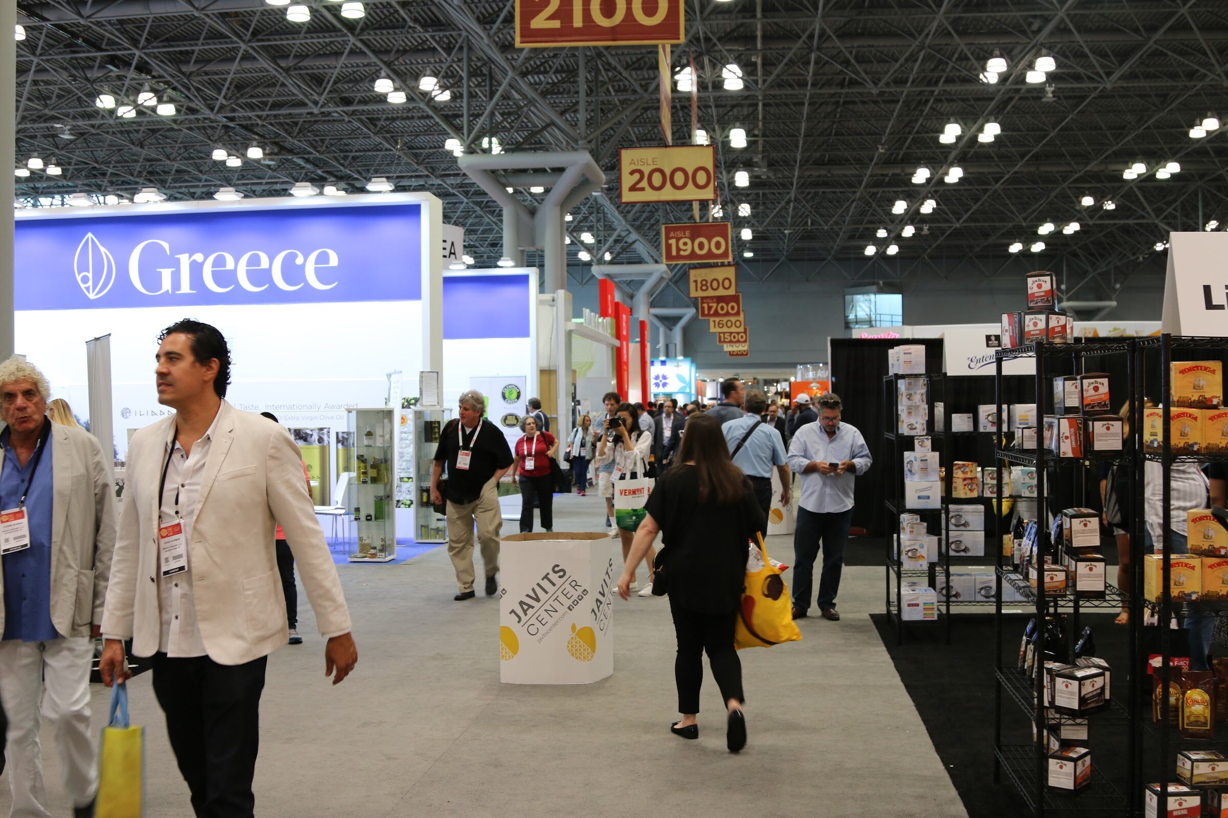 People with badges walking in a convention hall aisle with a blue Greece pavilion sign and aisle signs overhead.