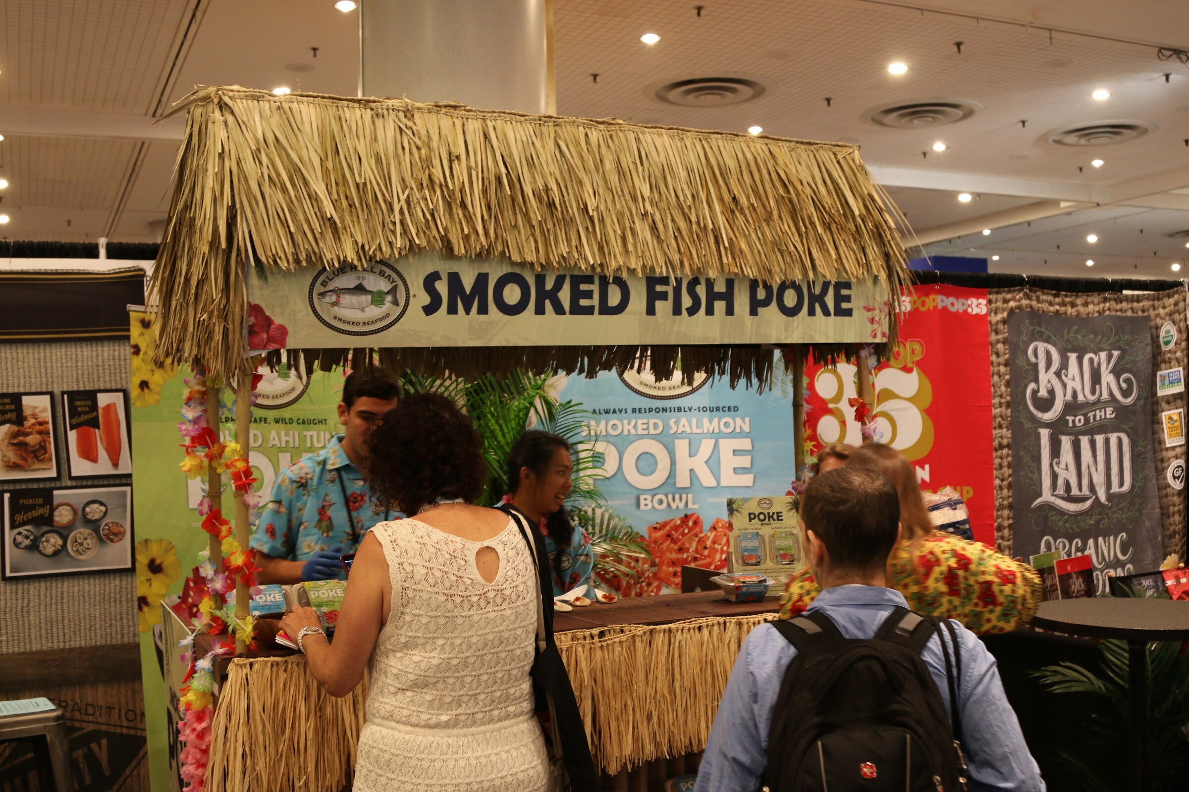 A booth with a straw roof and signs reading Smoked Fish Poke, with visitors in front and a Back to the Land banner at right.