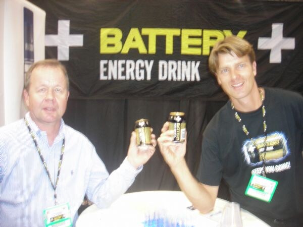 Two men with lanyards hold up cans of Battery Energy Drink in front of a black Battery Energy Drink banner.