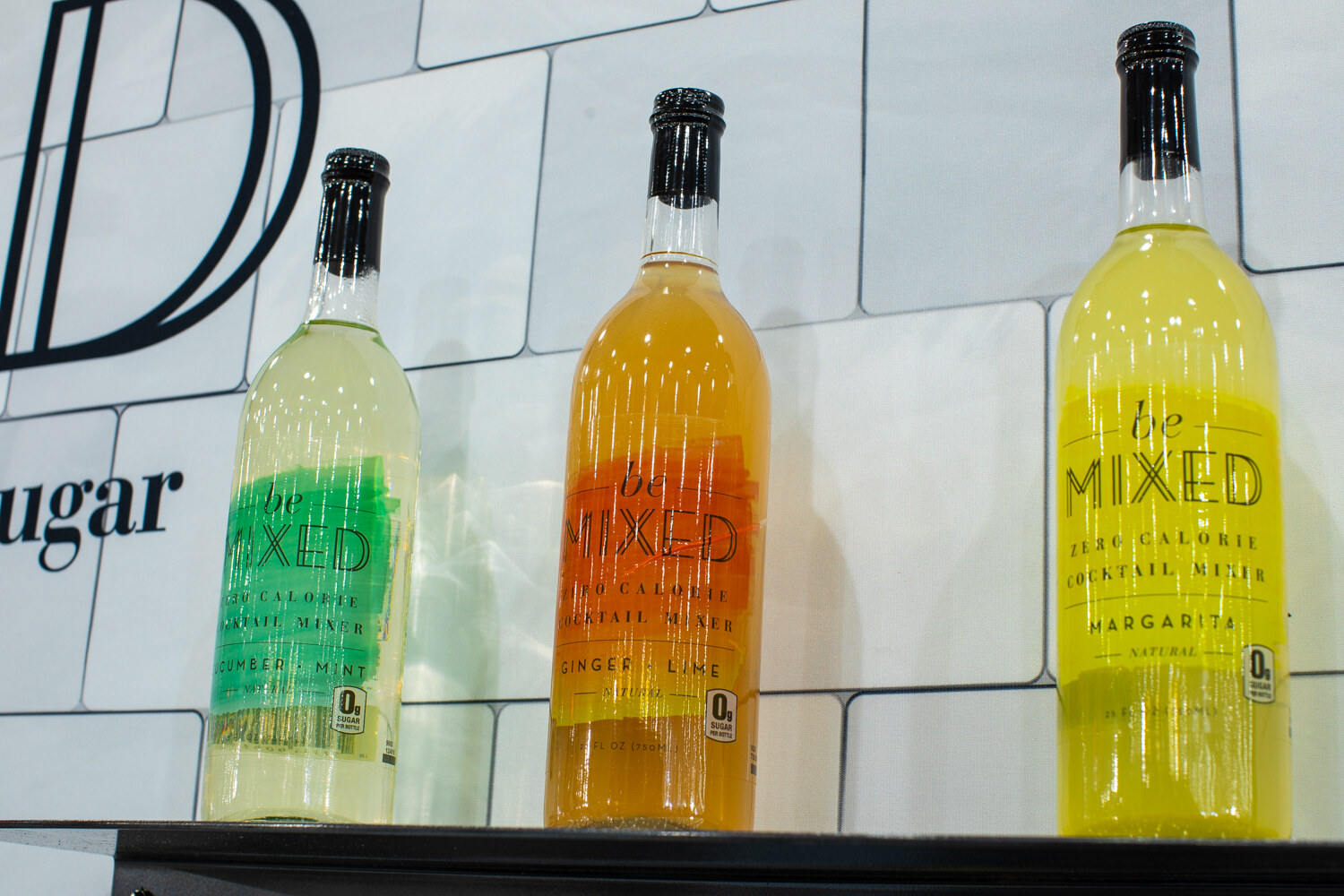 Three tall bottles of Be Mixed in green, orange and yellow on a white tile shelf.