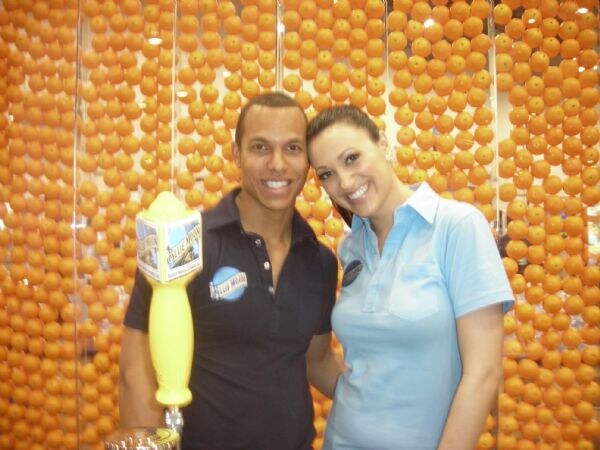 A man in a navy polo holding a yellow Blue Moon tap handle stands beside a woman in a light blue polo, in front of a wall of orange balls.