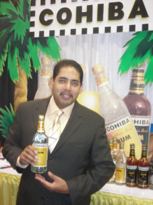 A man in a gray suit holds a bottle of Cohiba rum in front of a display with palm trees and large Cohiba rum bottle graphics.