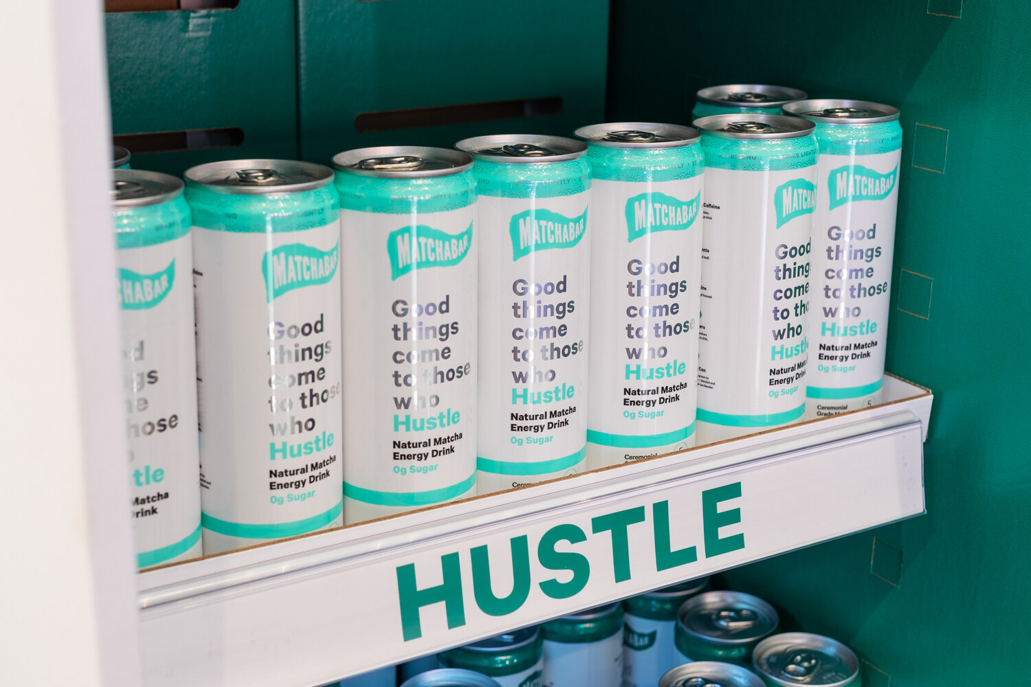 Cans of Hustle matcha energy drink on a teal shelf.