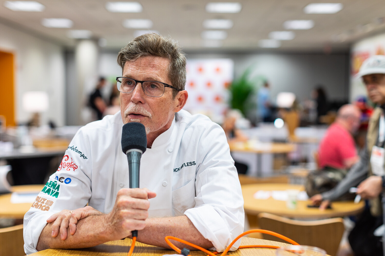 Man in a white chef's jacket speaking into a microphone with tables and people blurred behind him.