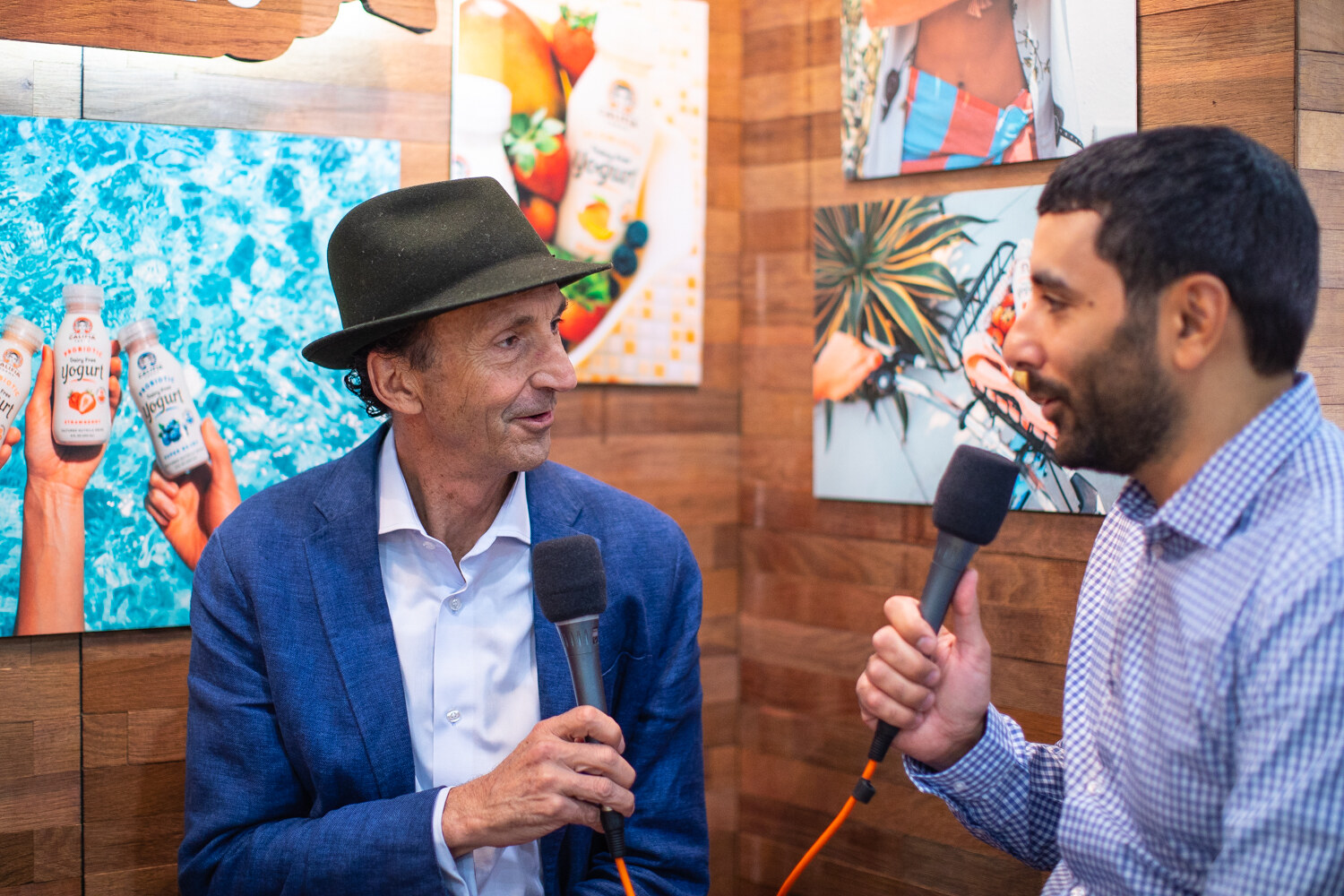 A man in a green hat and blue blazer and a man in a checked shirt holding microphones against a wood wall with tropical artwork.