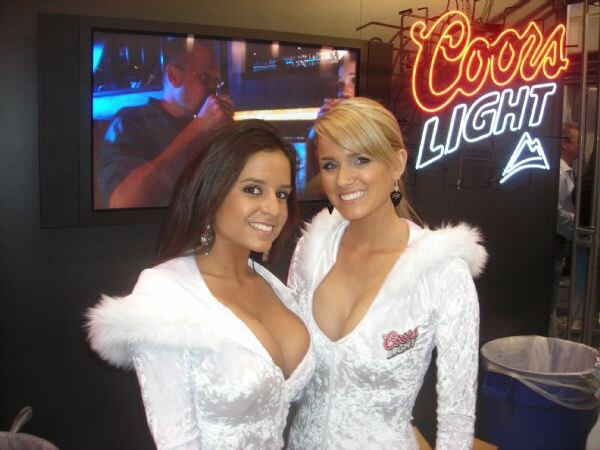 Two smiling women in white fur-trimmed velvet outfits with Coors Light logos stand in front of a lit Coors Light neon sign and a wall-mounted screen.