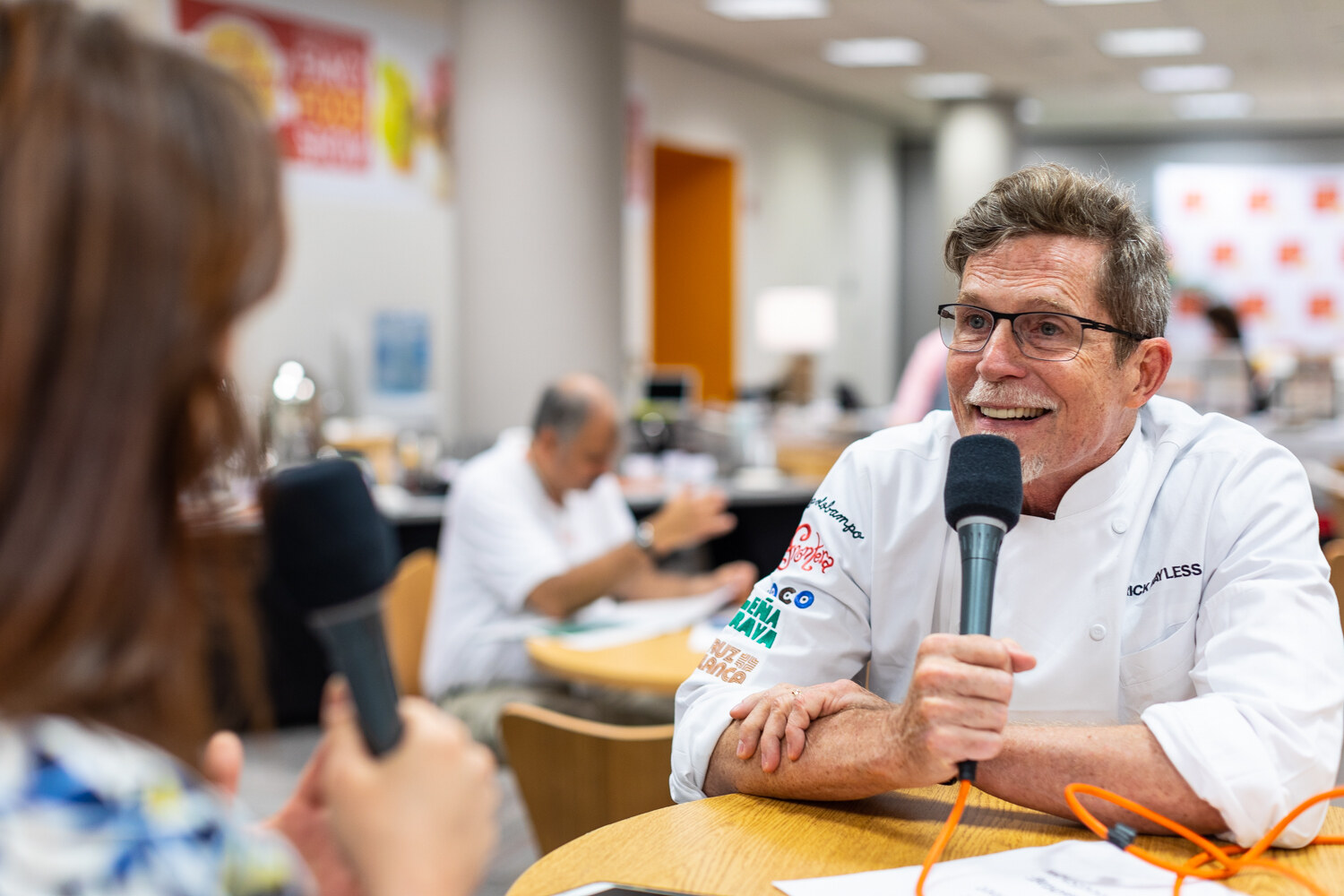 Man in a white chef's jacket with logo patches holding a microphone at a wooden table in a meeting room.