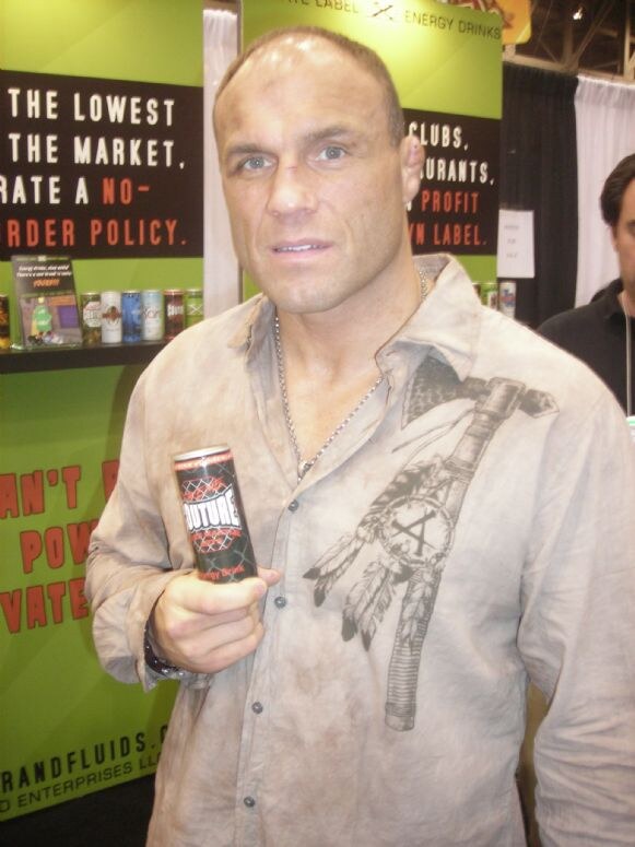 A bald man in a worn tan button shirt with a feather and tomahawk graphic holds a black Couture energy drink can in front of a green and black booth sign.