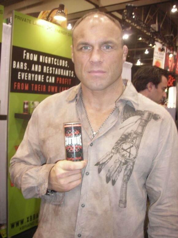 A bald man in a worn tan shirt with a feather graphic holds a Couture energy drink can in front of a green booth sign about nightclubs, bars and restaurants. Another man stands behind him.