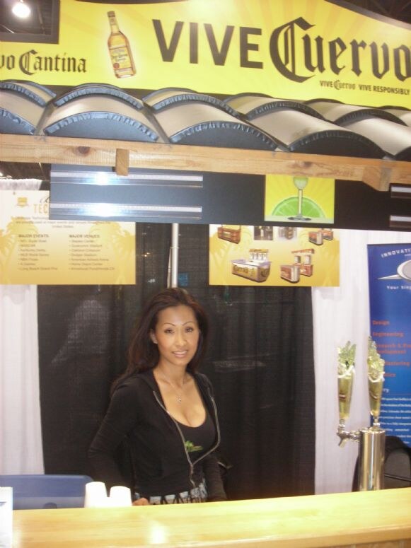 A woman in a black zip jacket stands behind a wooden bar counter under a yellow "Vive Cuervo" banner, with a tap tower to her right and display panels behind her.