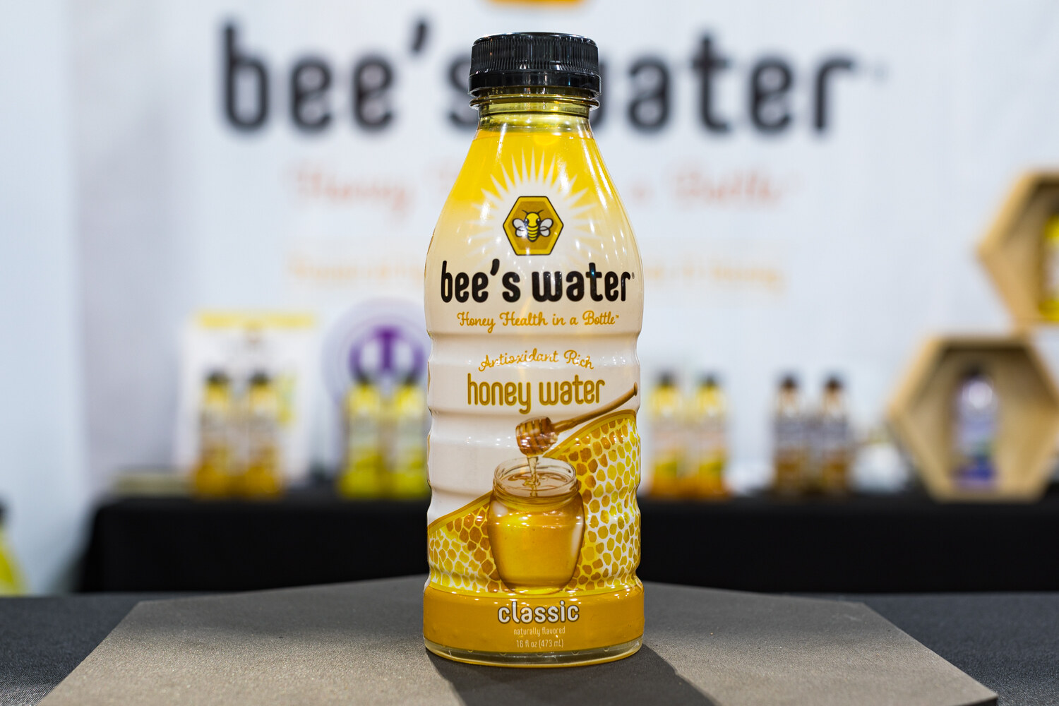 Photo of a single bottle of bee's water honey water in front of a blurred booth sign.