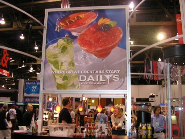 A large Daily's Cocktails & Mixers sign showing two cocktails hangs above a booth where several people stand behind a counter holding bottles and ice.