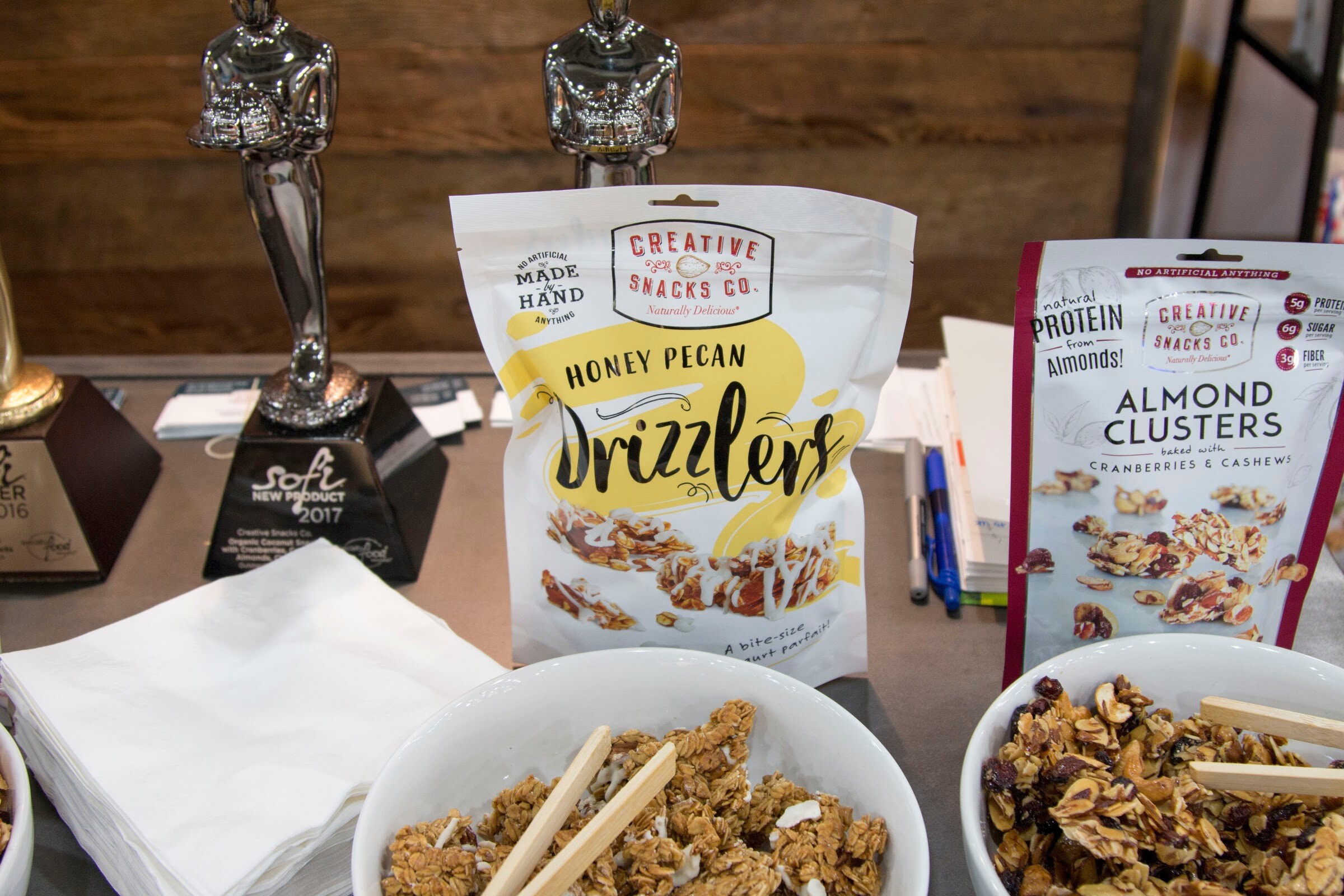 Photo of Creative Snacks Drizzlers bags and bowls of granola beside trophies.
