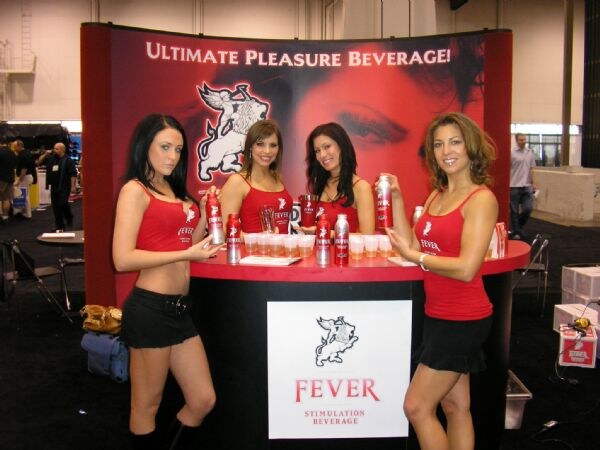 Four women in red tops stand behind a round Fever counter holding cans of the drink, with a red banner reading "Ultimate Pleasure Beverage!" behind them.