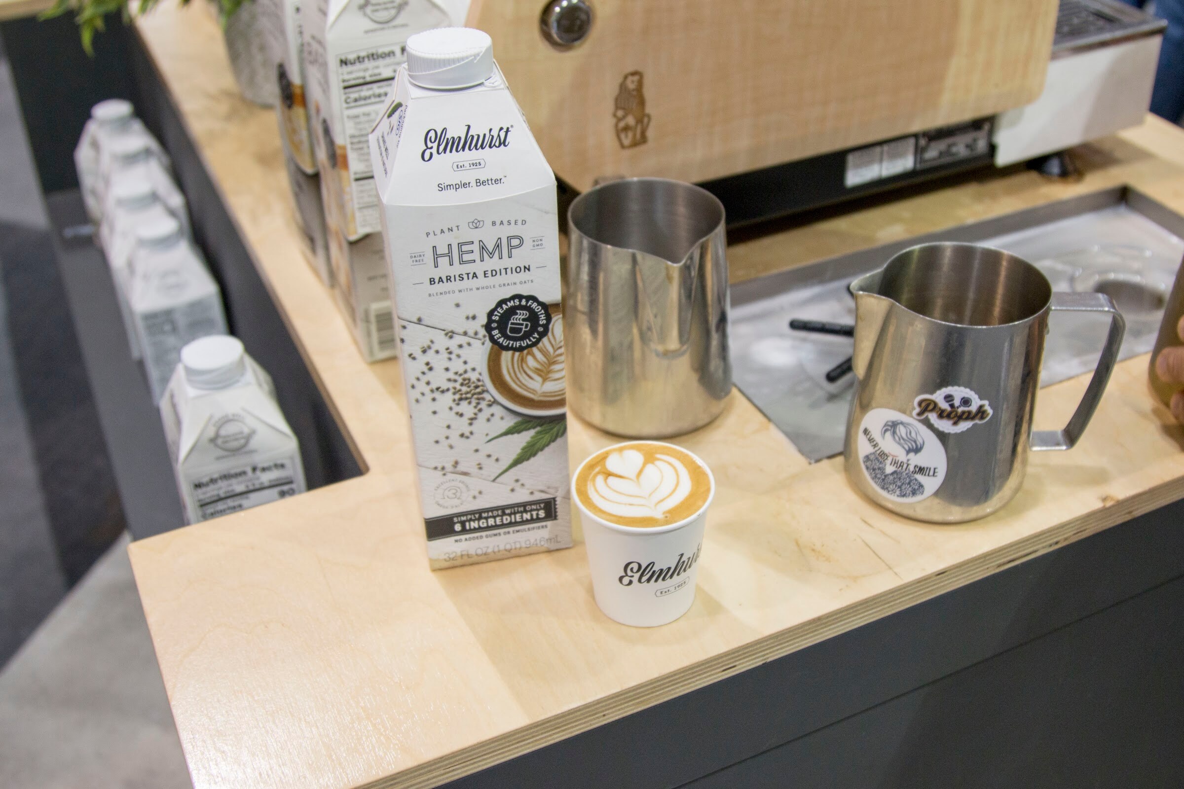 Photo of a carton of Elmhurst hemp barista milk beside a latte and steaming pitchers.