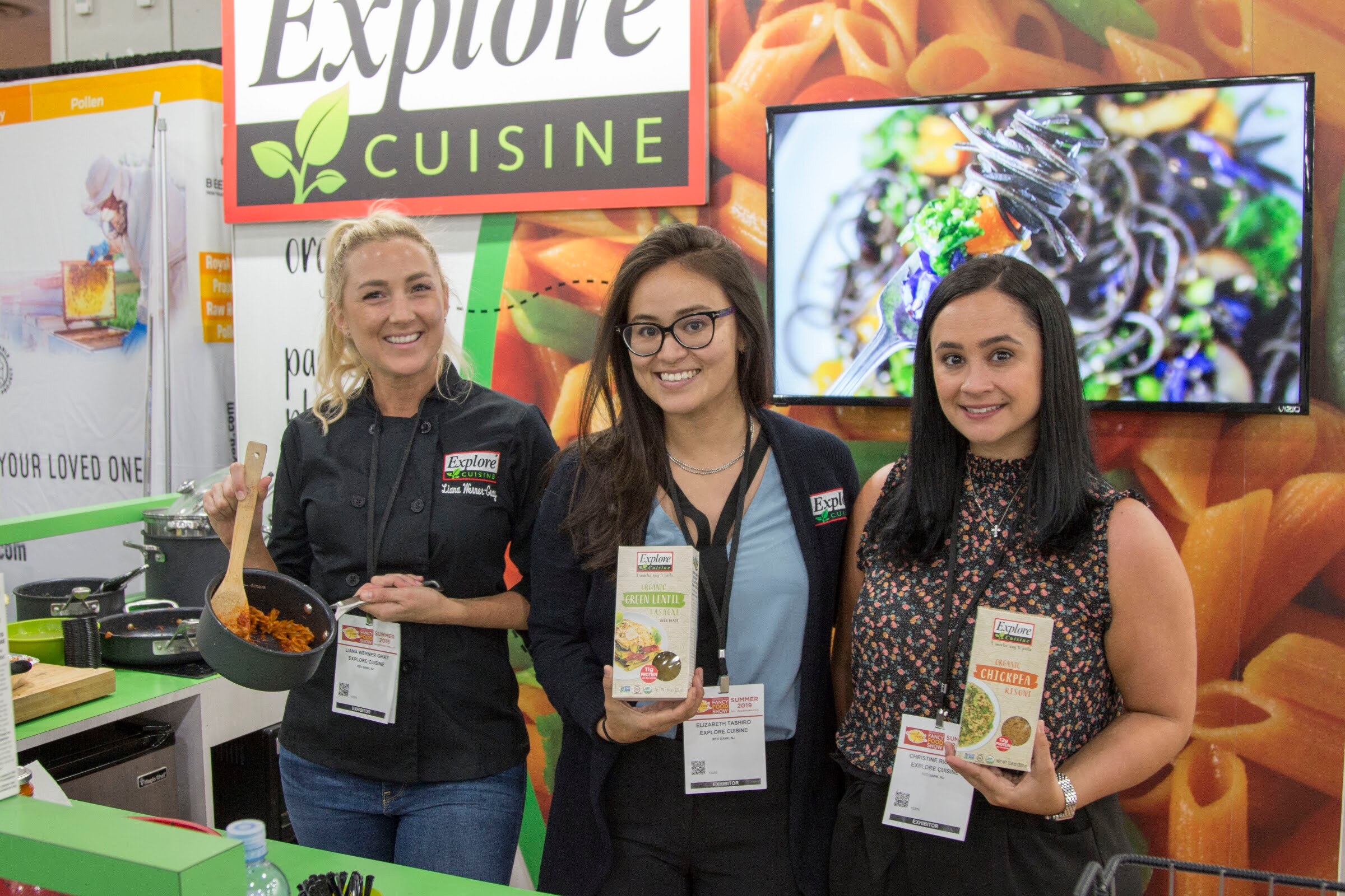 Photo of three women in front of an Explore Cuisine booth, two holding product boxes.