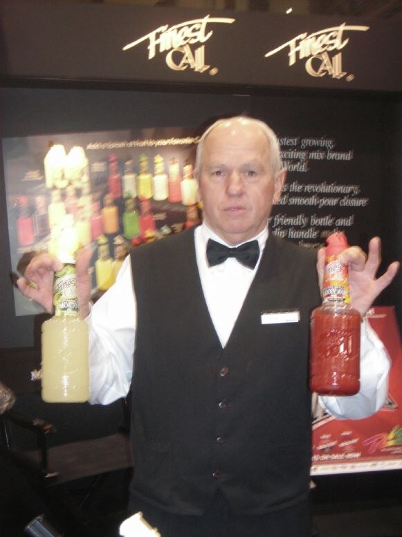 A man in a black vest and bow tie holds up a bottle of Finest Call mixer in each hand in front of Finest Call signs and a display of mixer bottles.