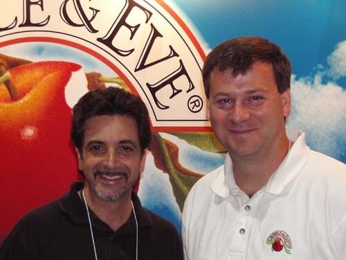 Two smiling men pose close to the camera in front of a large Apple & Eve sign showing a red apple; the man on the right wears a white polo with an Apple & Eve logo.