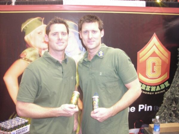 Two men in green polo shirts each hold a can of Grenade Energy Drink in front of a Grenade banner showing a military-style logo.