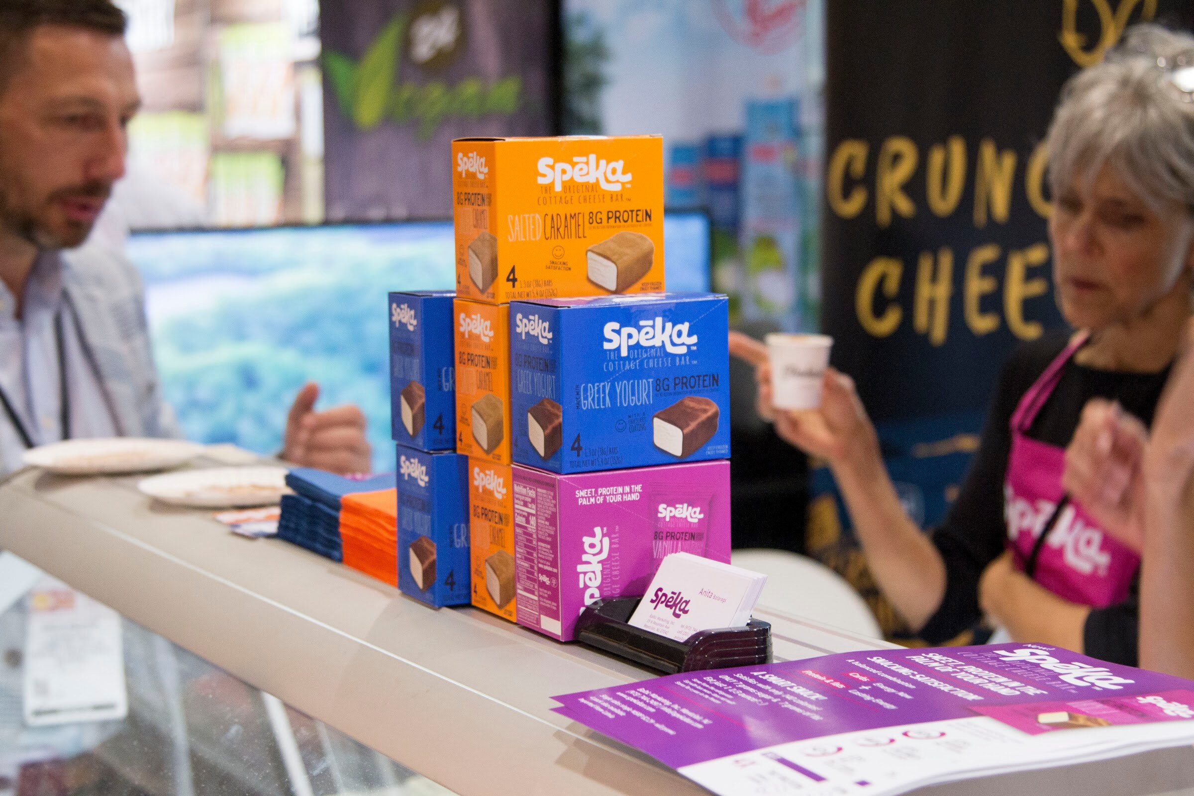 Photo of stacked boxes of protein snacks with two exhibitors sampling at the booth.