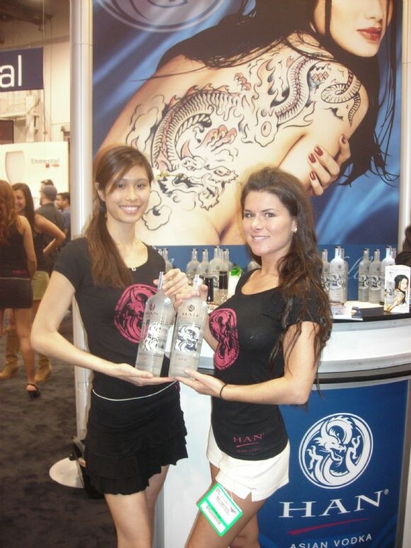 Two women in black Han T-shirts hold bottles of Han Asian Vodka at a booth with a dragon-tattoo poster and a Han logo on the counter.