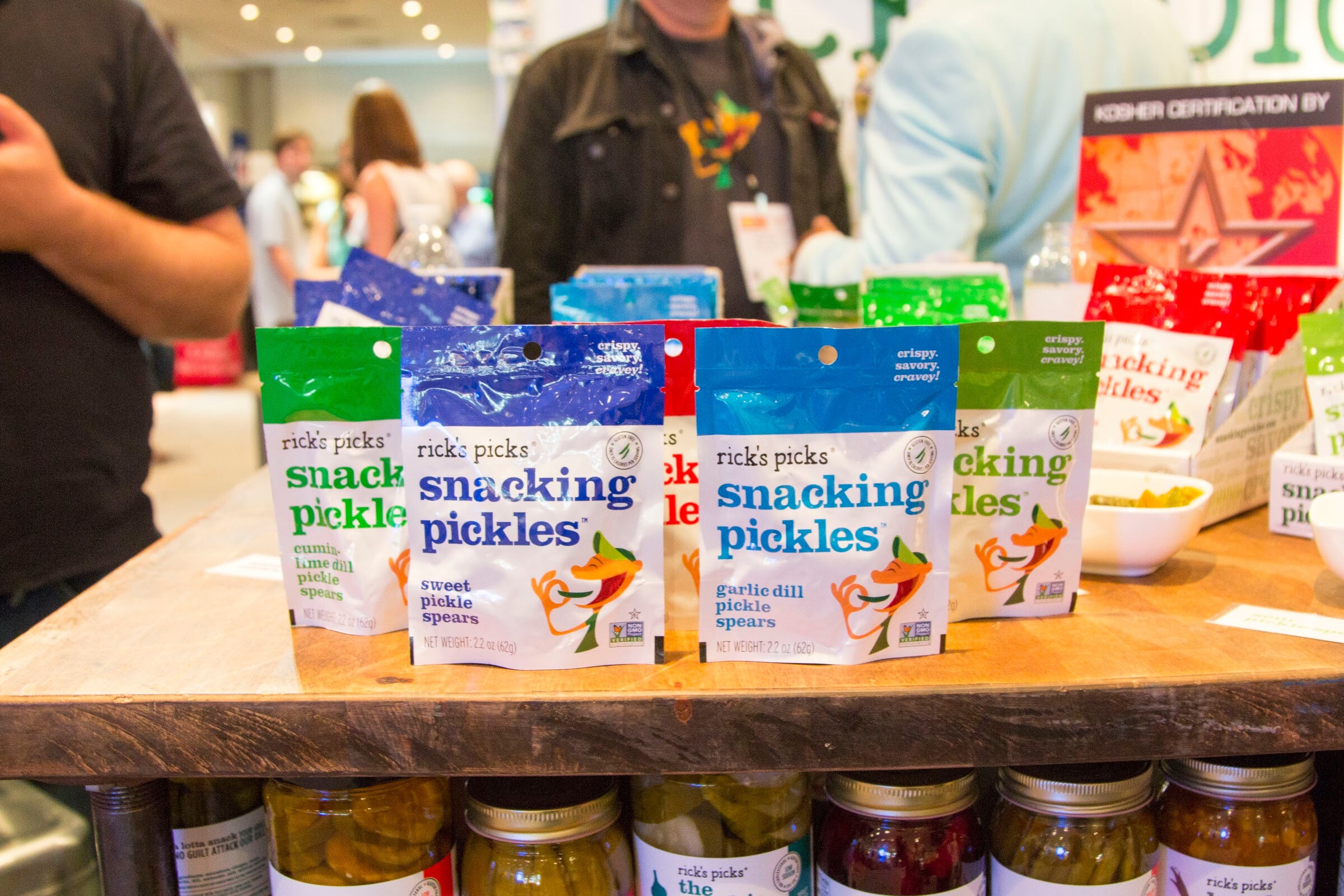 Photo of bags of Rick's Picks snacking pickles on a table with pickle jars.