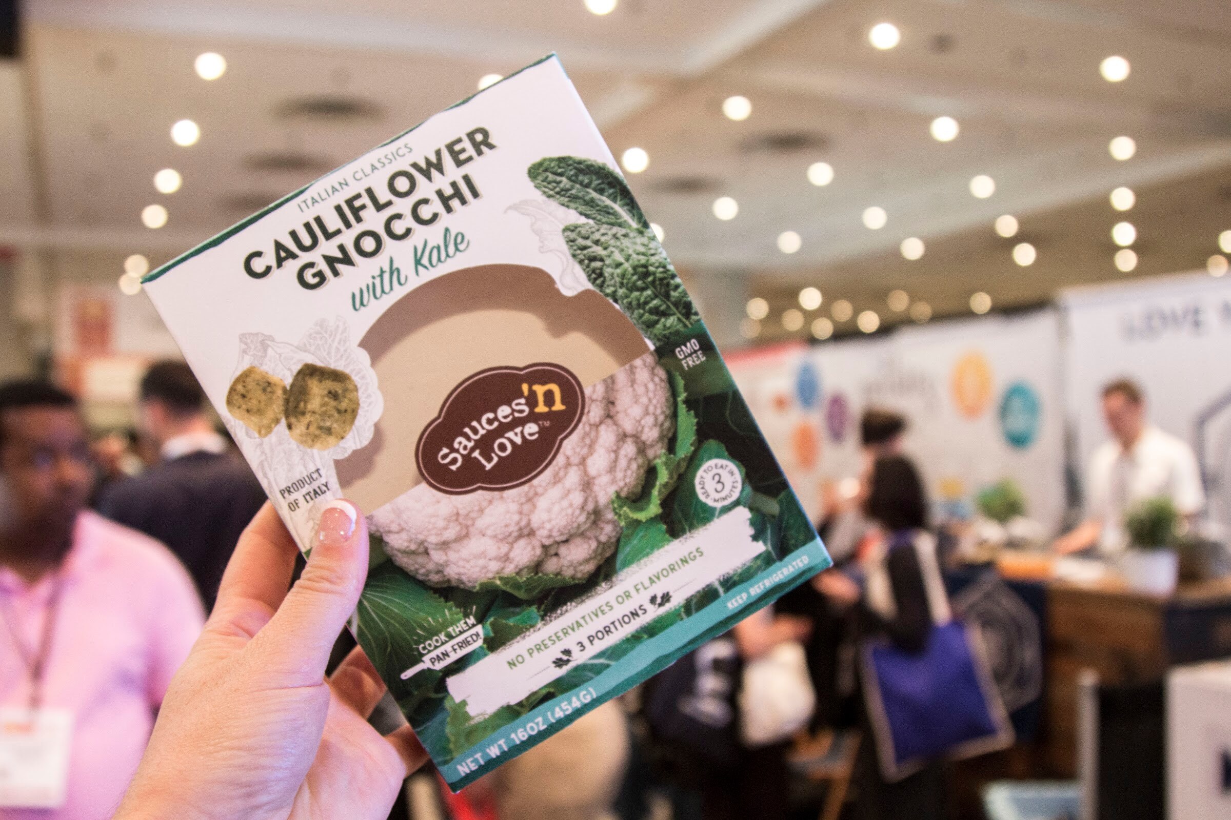 Photo of a hand holding a Sauces 'n Love cauliflower gnocchi box with the show floor behind.