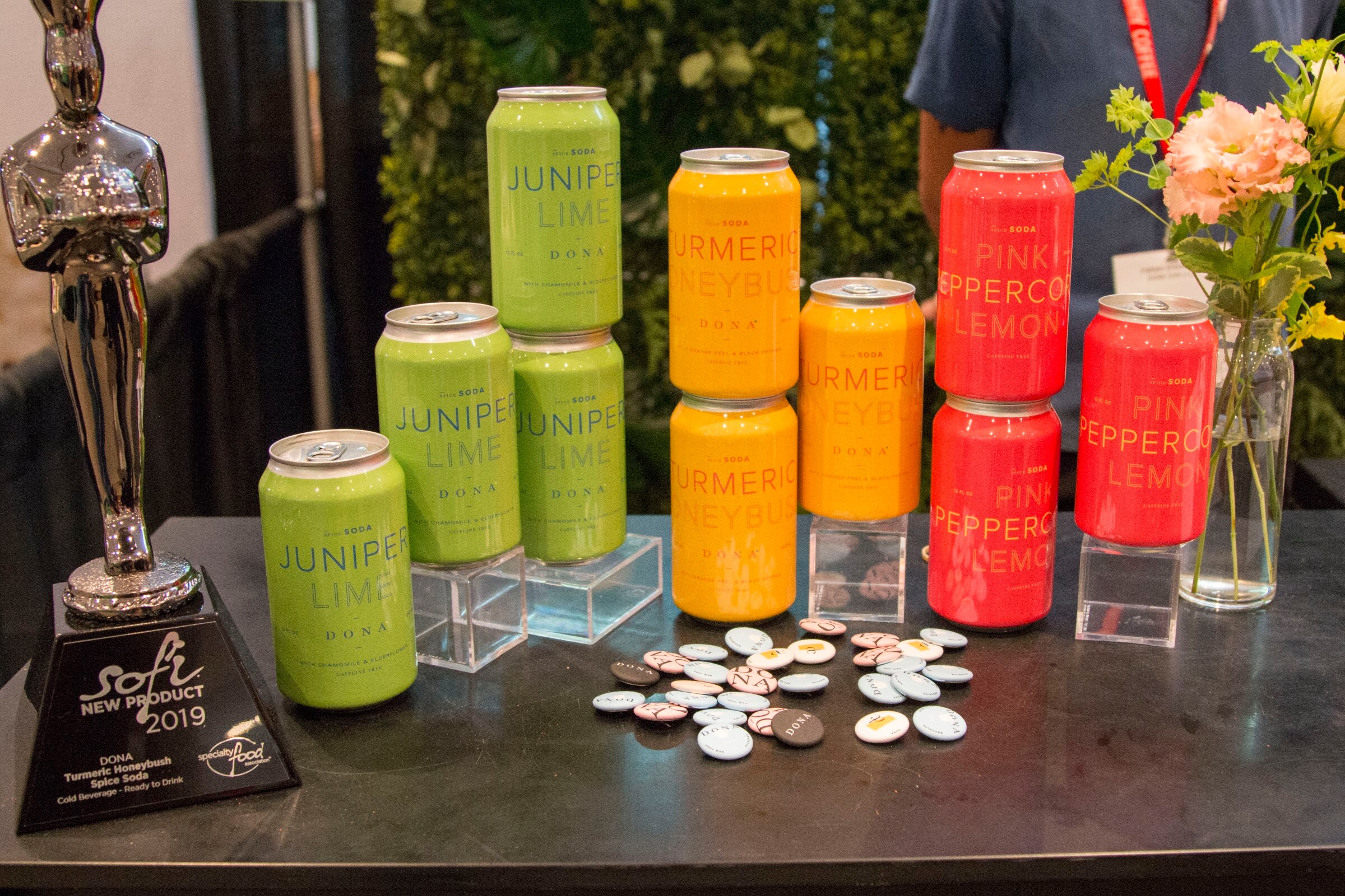 Photo of cans of sparkling soda in lime, turmeric ginger and pink pepper lemon beside a 2019 sofi new product award trophy.