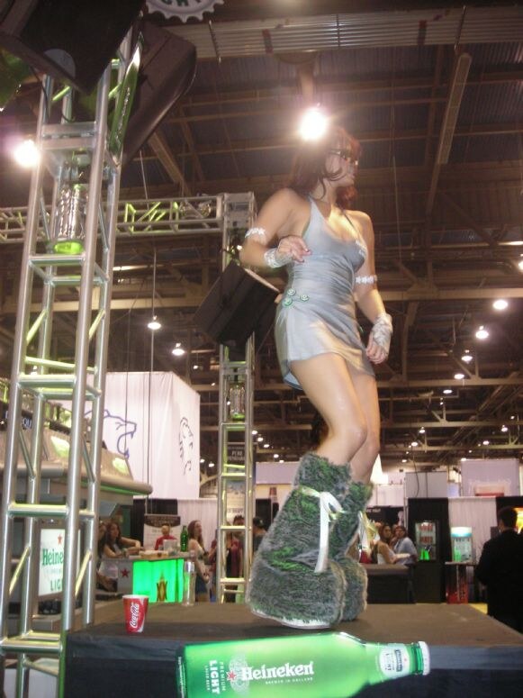 A woman in a gray dress and furry boots dances on a platform beside metal truss. A Heineken bottle lies in the foreground, and a Coca-Cola cup sits nearby.