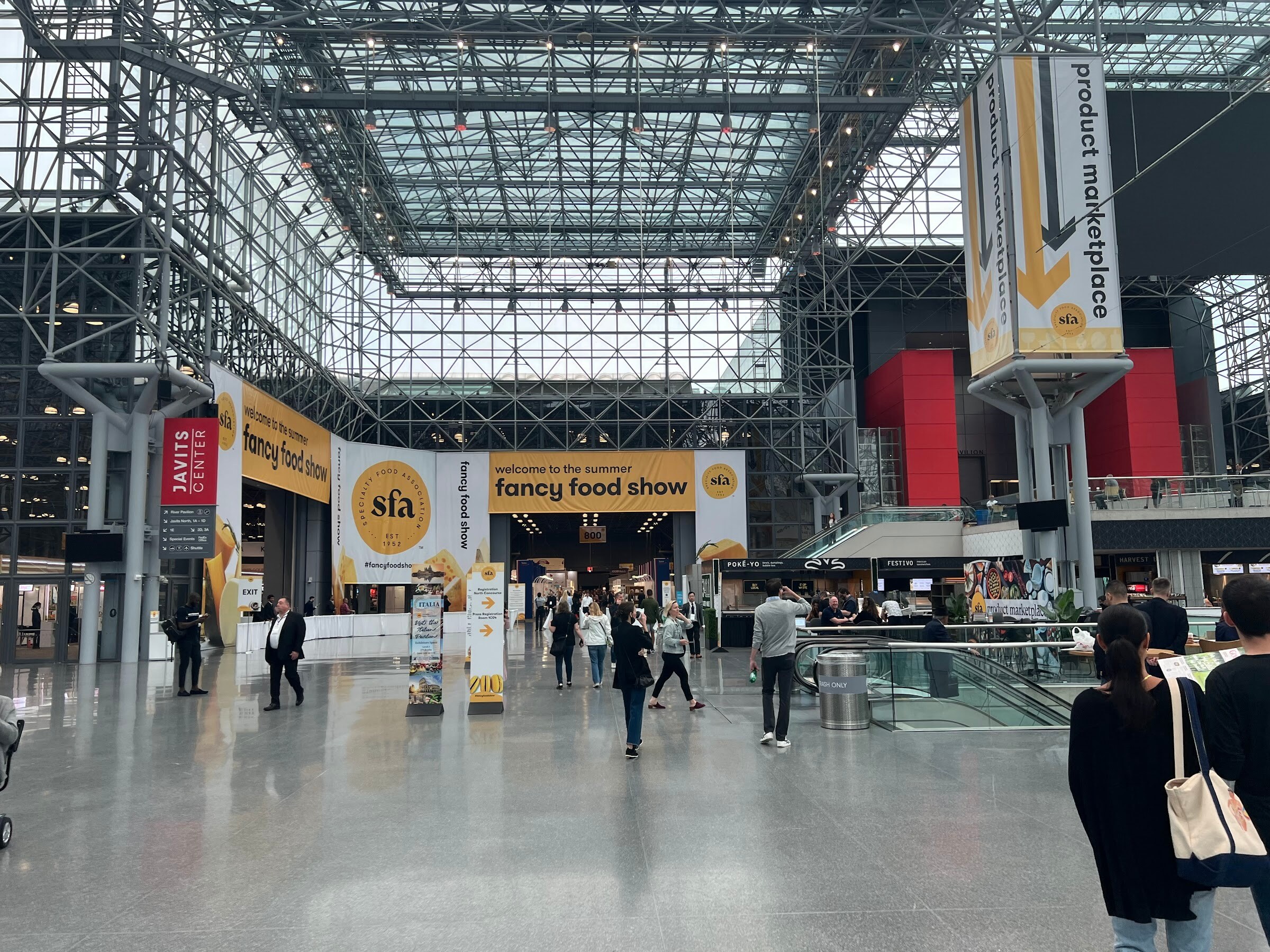 Visitors walk through the glass-roofed Javits Center atrium under fancy food show and product marketplace banners