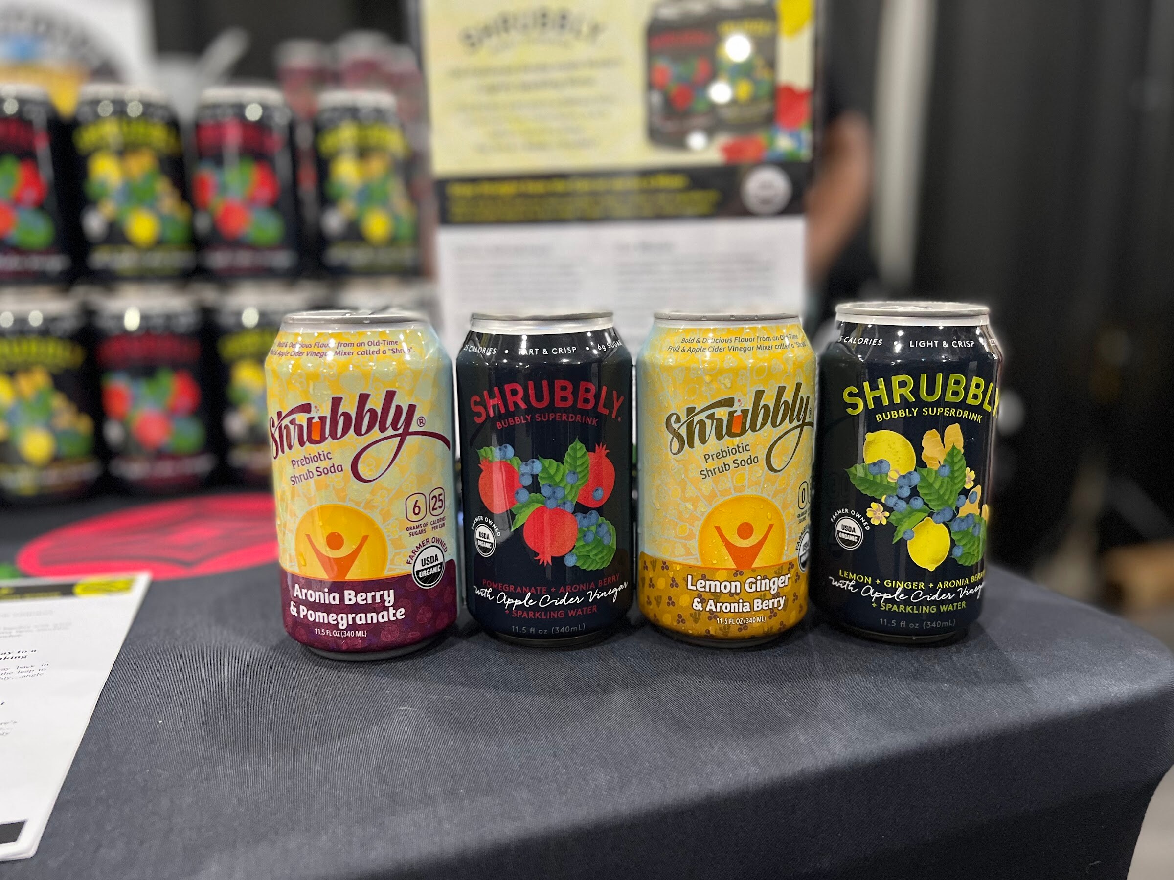 Four Shrubbly cans in aronia berry pomegranate, apple cider vinegar and lemon ginger flavors stand in front of stacked cans