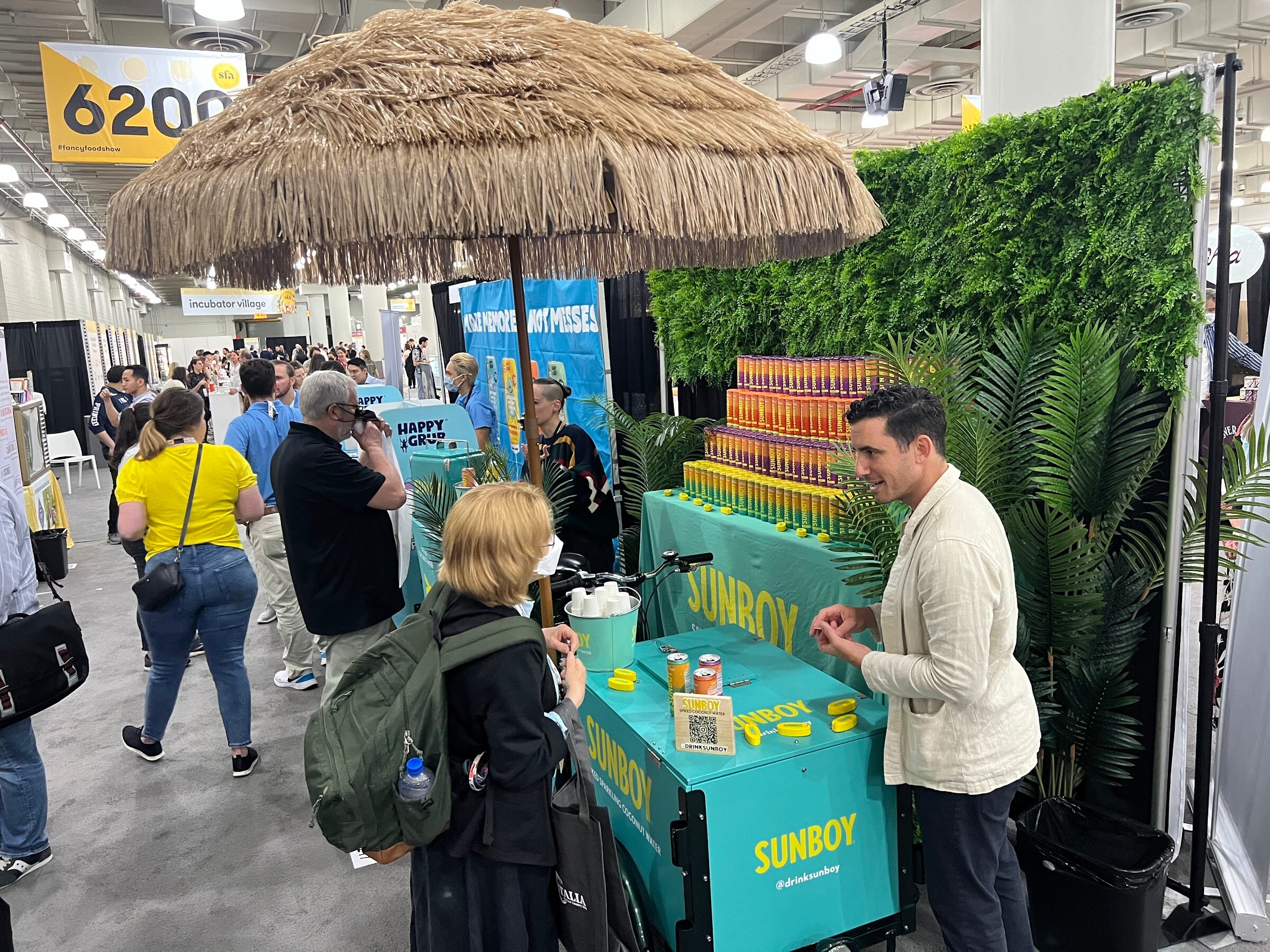 Visitors stop at a teal Sunboy cart under a thatched umbrella with stacked cans and tropical plants in a busy aisle