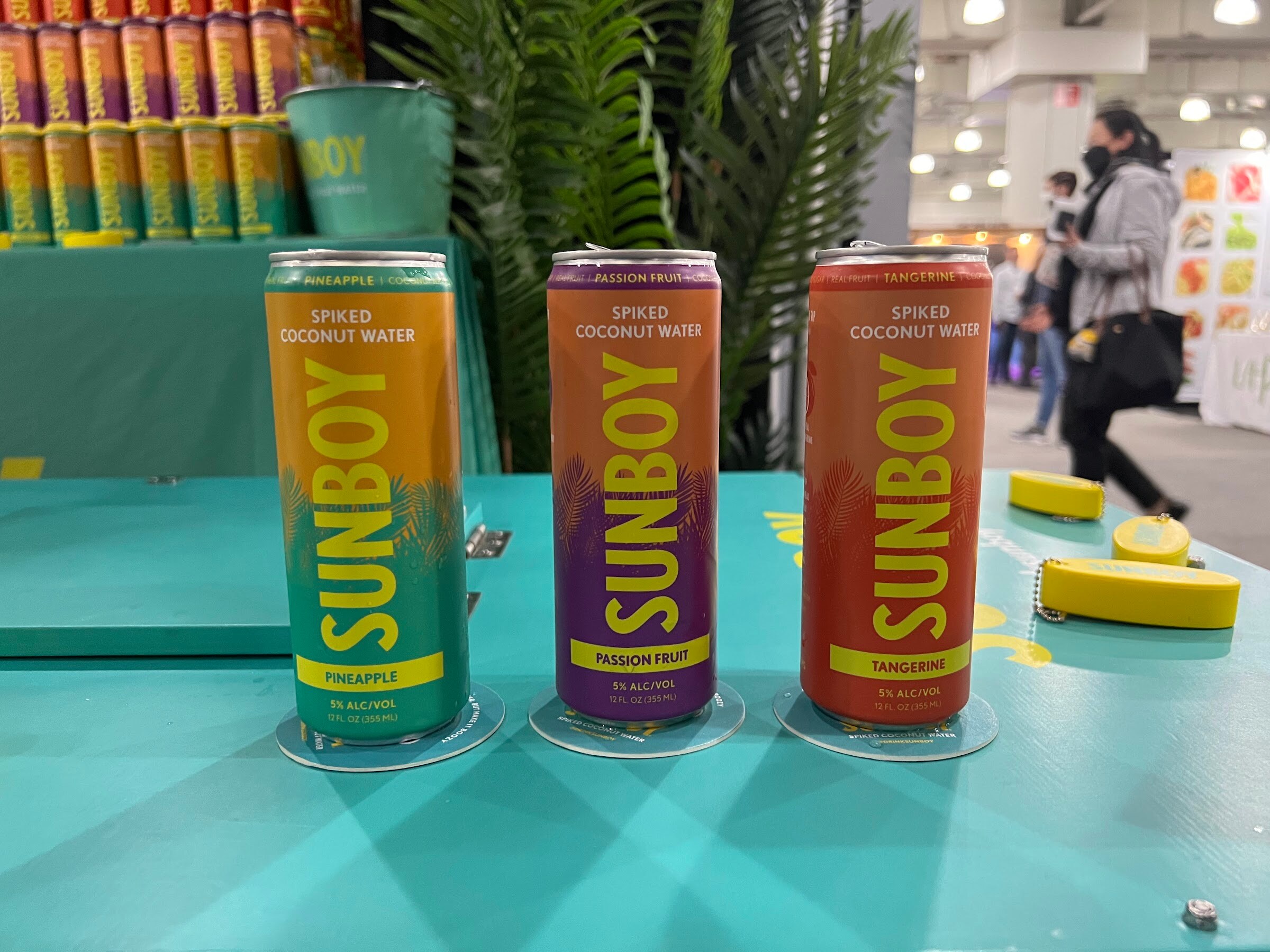 Three Sunboy spiked coconut water cans in pineapple, passion fruit and tangerine stand on a teal table