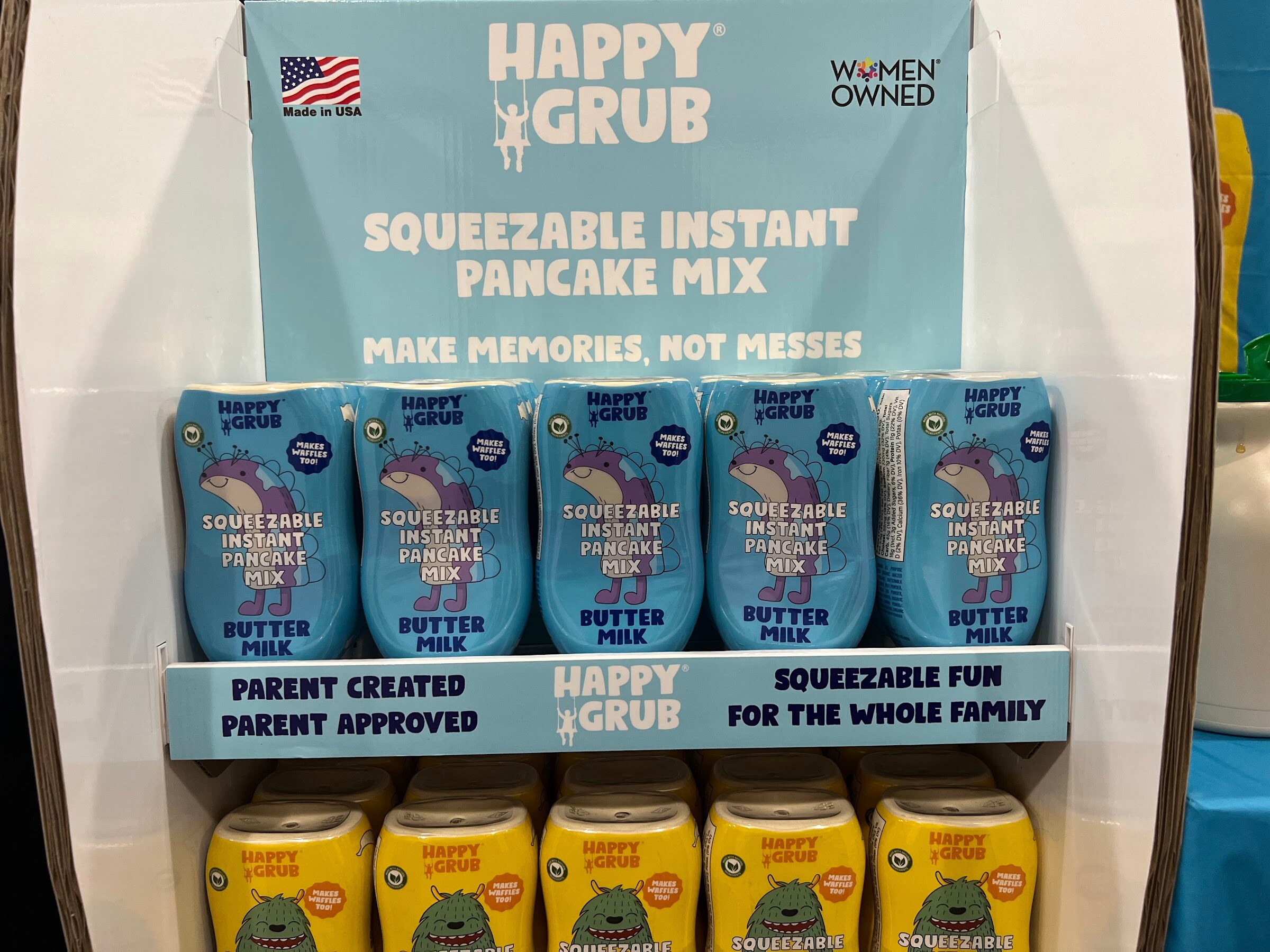 Blue Happy Grub squeezable instant pancake mix bottles are stacked on a display under a Women Owned banner
