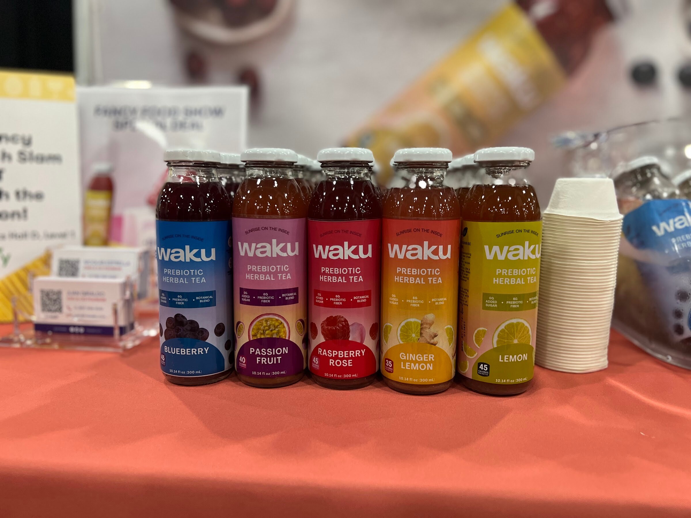 Five waku prebiotic herbal tea bottles in blueberry, passion fruit, raspberry rose, ginger lemon and lemon stand on an orange tablecloth