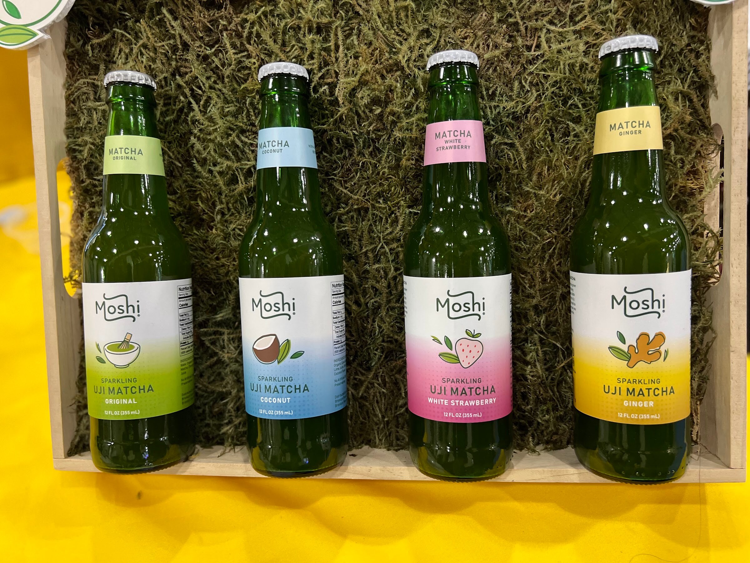 Four Moshi sparkling uji matcha bottles in original, coconut, white strawberry and ginger sit in a wooden crate on moss