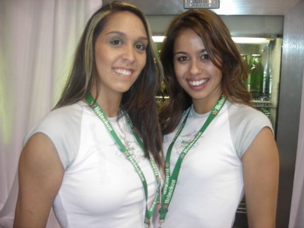 Two smiling women with long brown hair wear white t-shirts and green Heineken lanyards. Behind them is a lit refrigerated display holding green bottles, with white curtains at left.