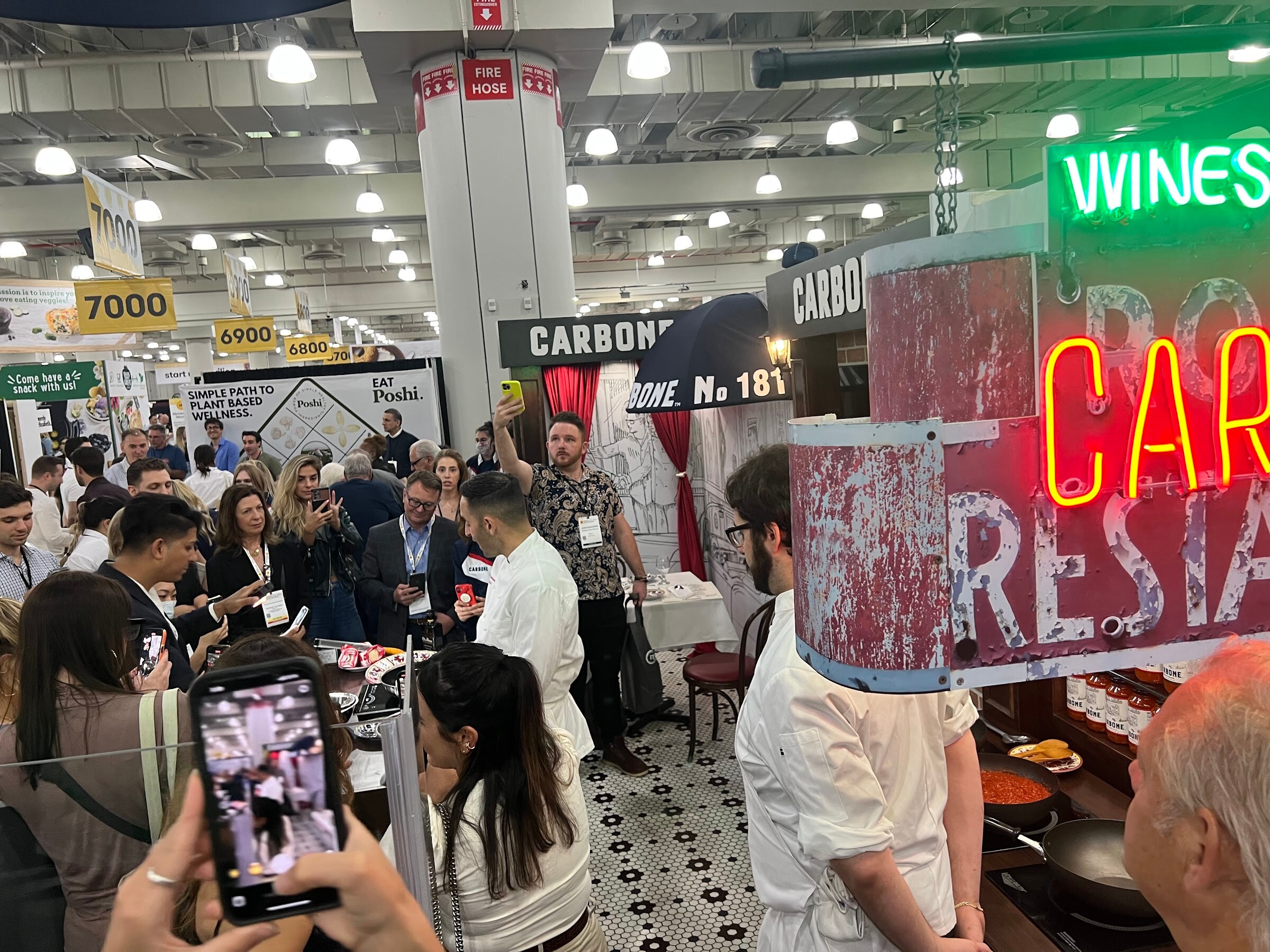 A crowd with phones raised watches chefs cook at a booth with a neon sign and black-and-white tiled floor
