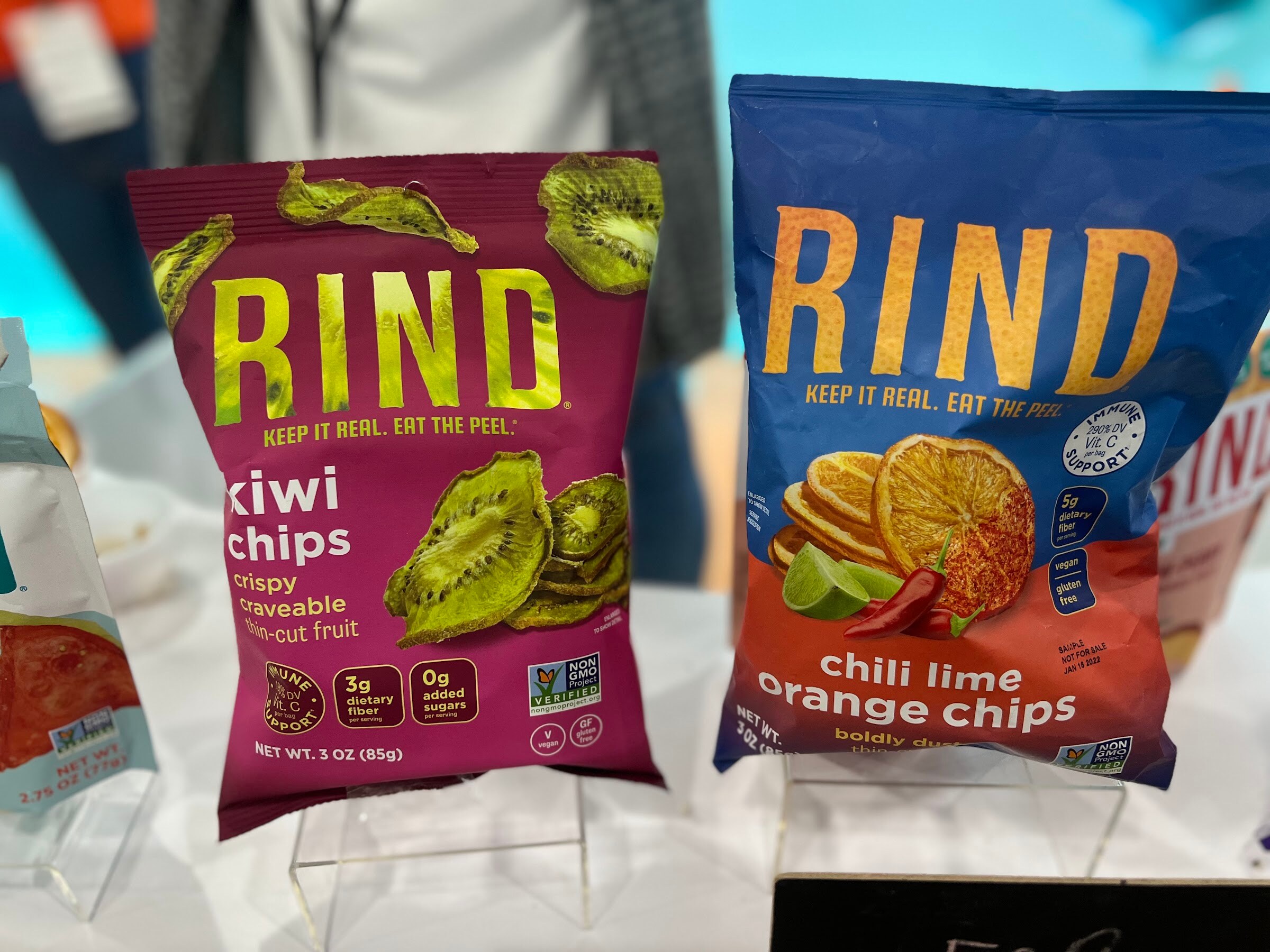 RIND kiwi chips and chili lime orange chips bags stand on a table in front of other RIND snacks