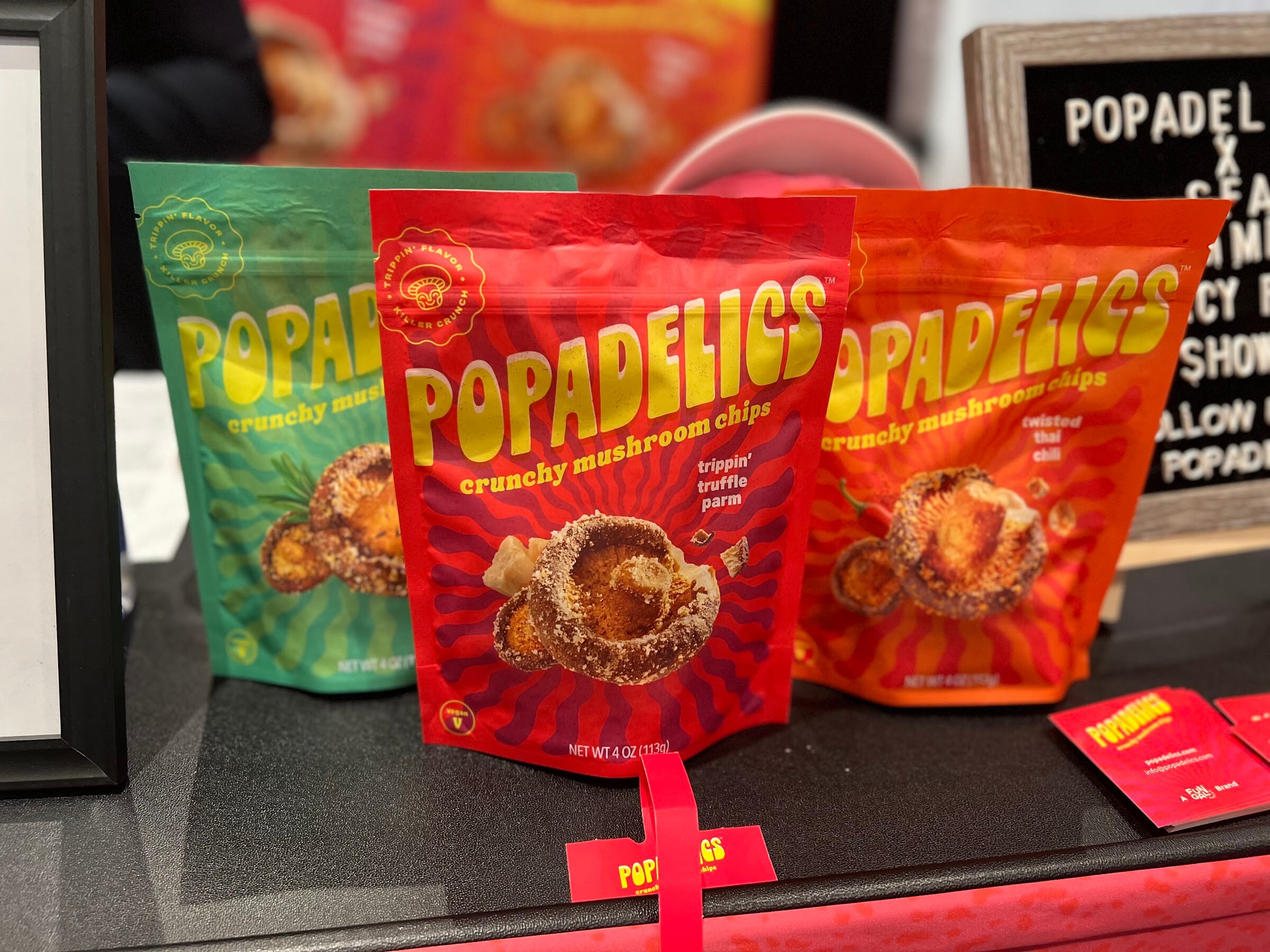 Colorful Popadelics crunchy mushroom chips bags stand on a table with a Popadelics sign behind