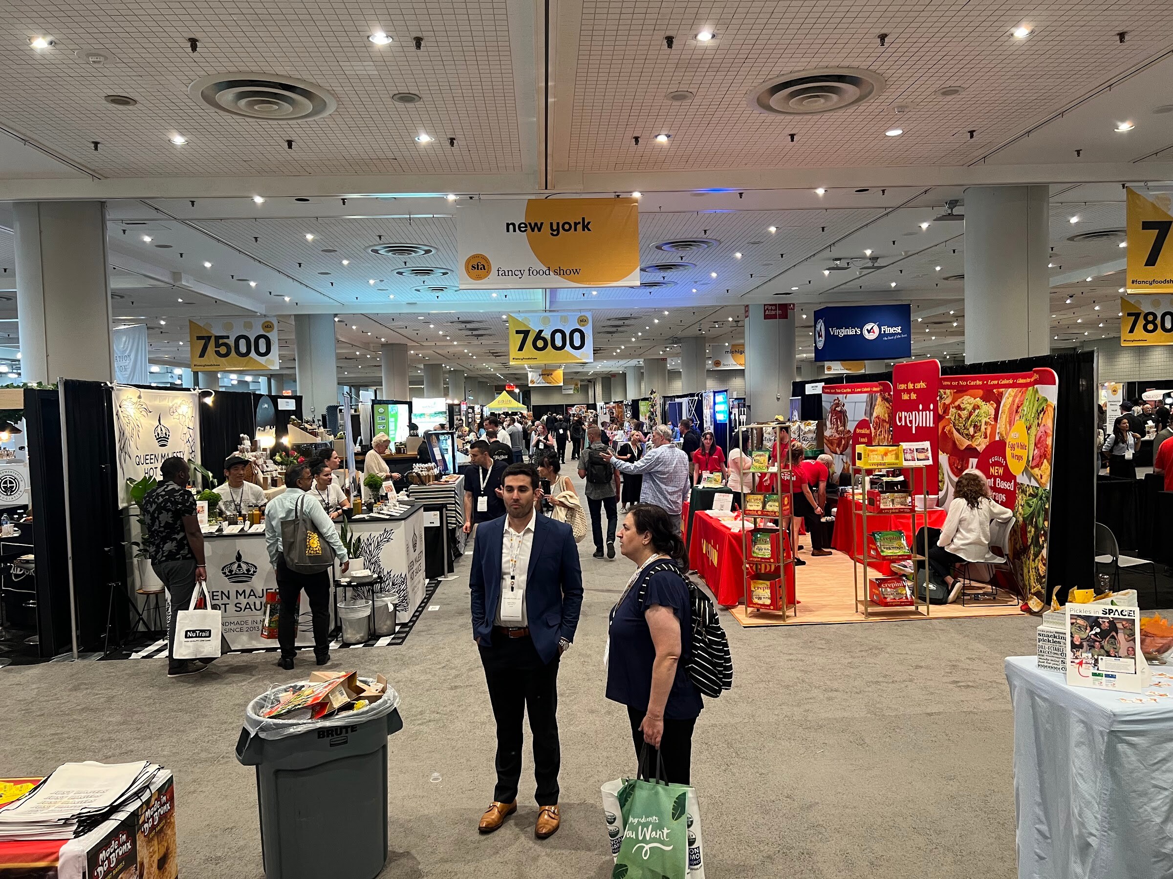 Two attendees stand in an aisle of the exhibit hall under a new york hanging sign with numbered booth aisles behind them