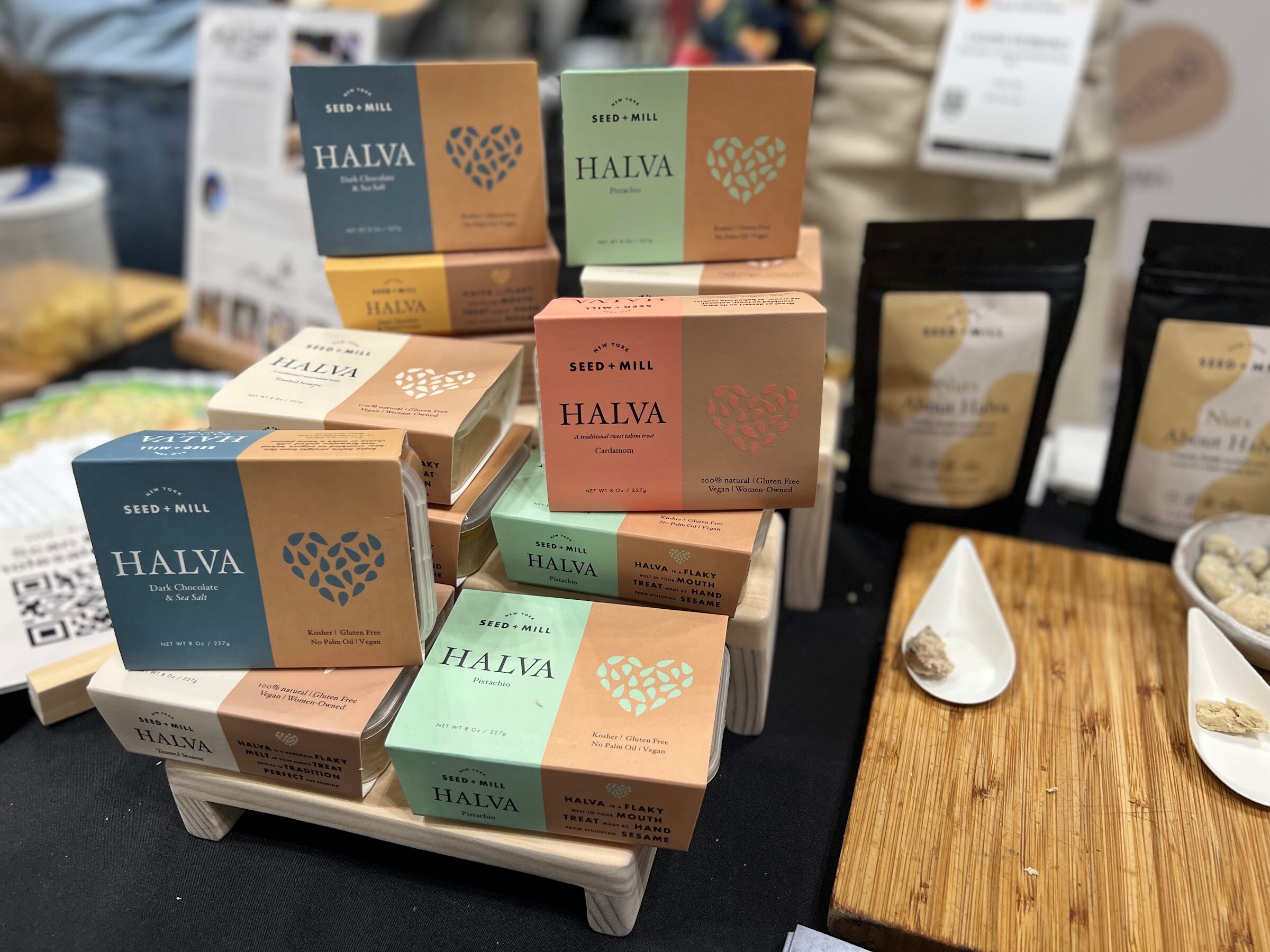 Boxes of Seed and Mill halva in blue, green and orange stand beside a wooden serving board with a spoon of halva