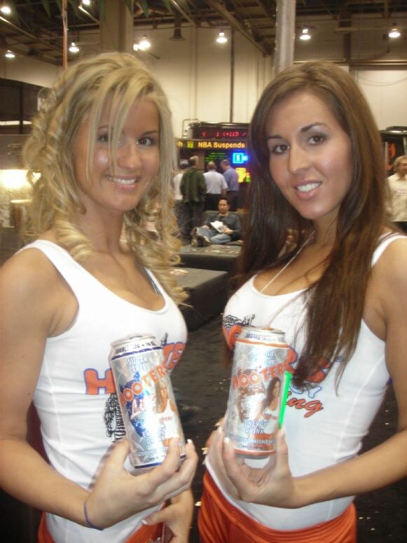 Two smiling women in white Hooters tank tops each hold a silver can of Hooters Lite energy drink. One has curly blonde hair and the other long brown hair; the show floor is behind them.