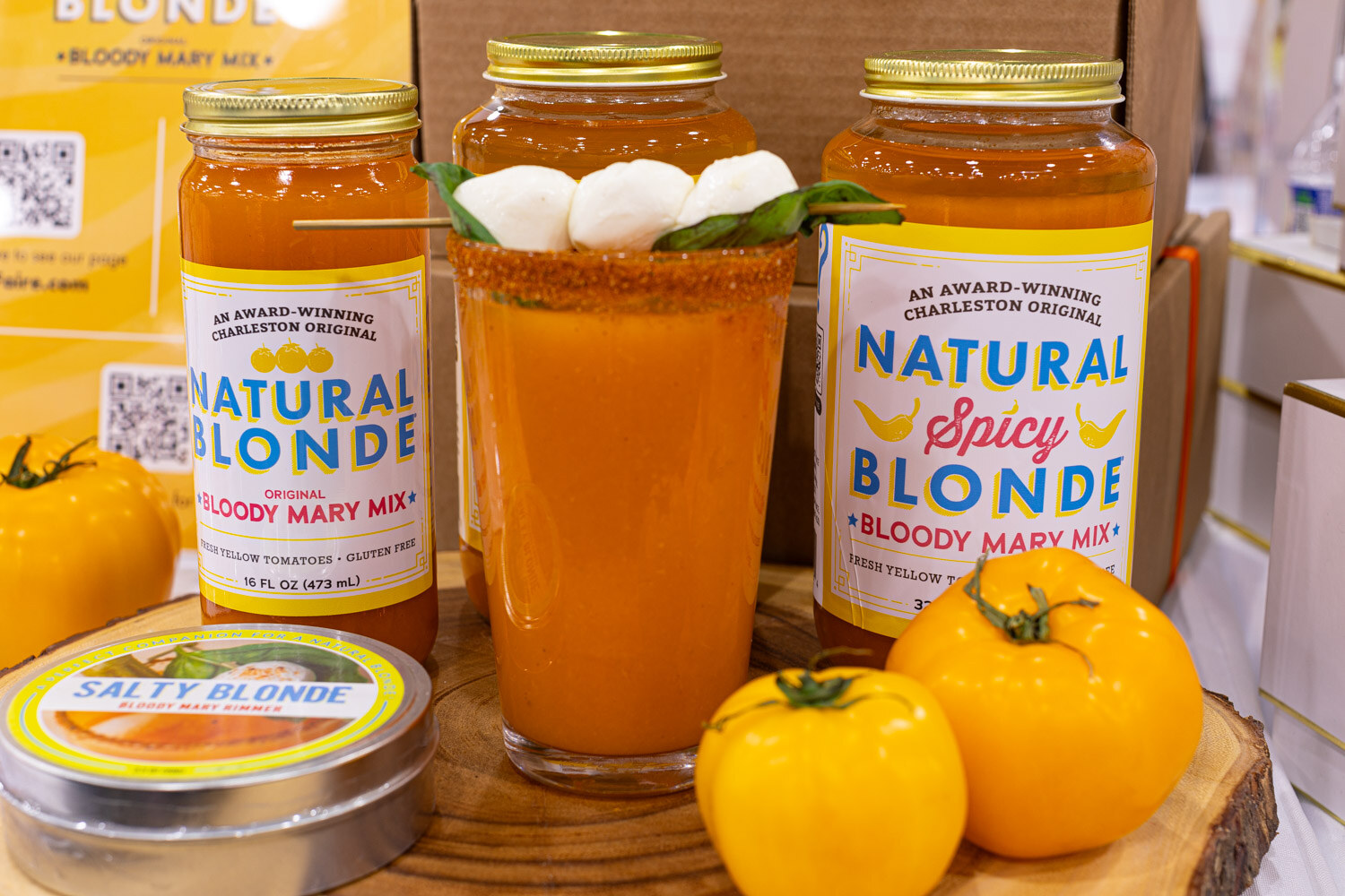 Jars of Natural Blonde Natural Spicy Blonde Bloody Mary Mix flank a garnished cocktail on a wooden slab with yellow tomatoes
