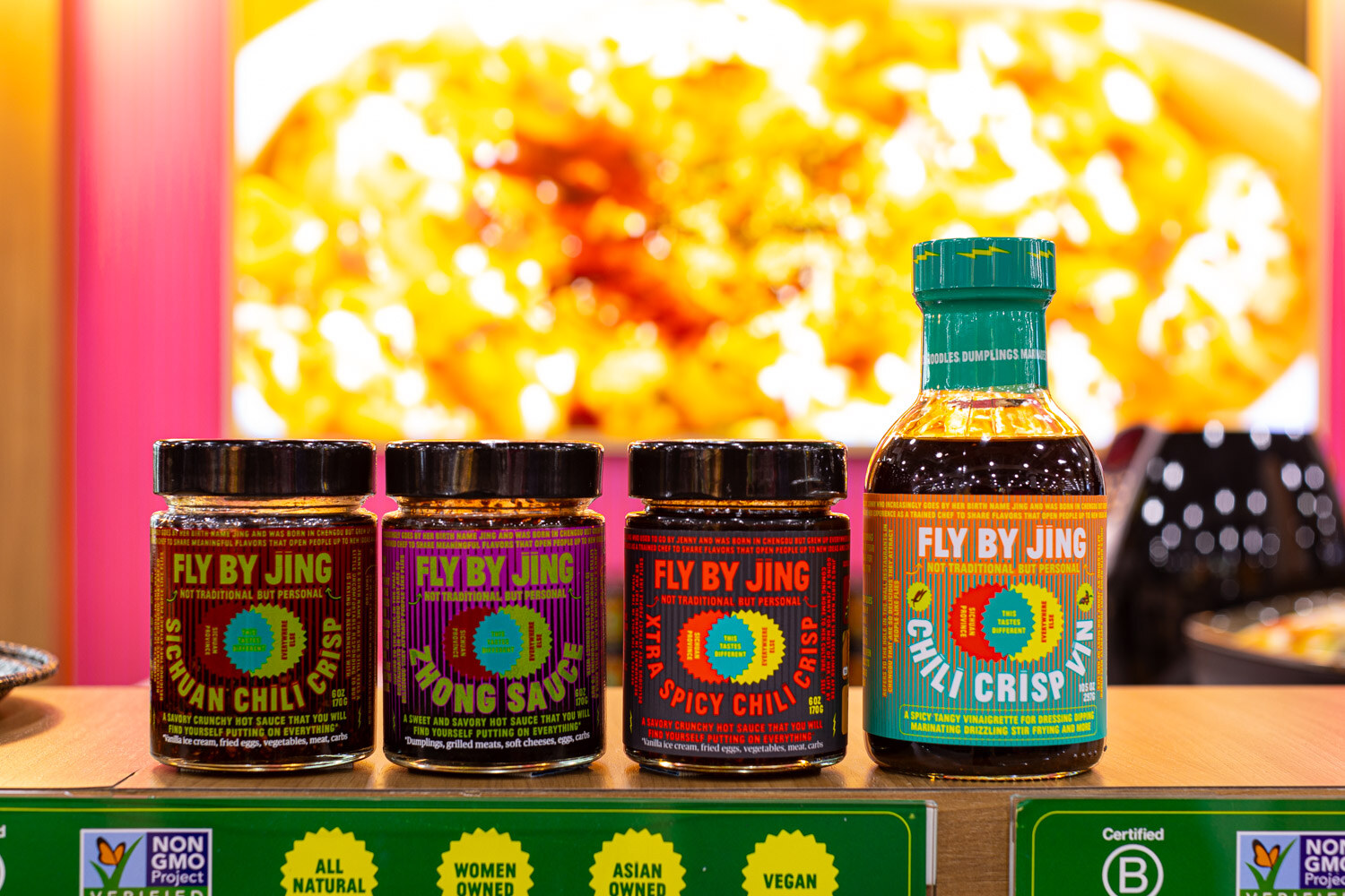 Jars and bottles of Fly By Jing Sichuan chili crisp and sauces sit on a shelf in front of a bright pink and yellow backdrop