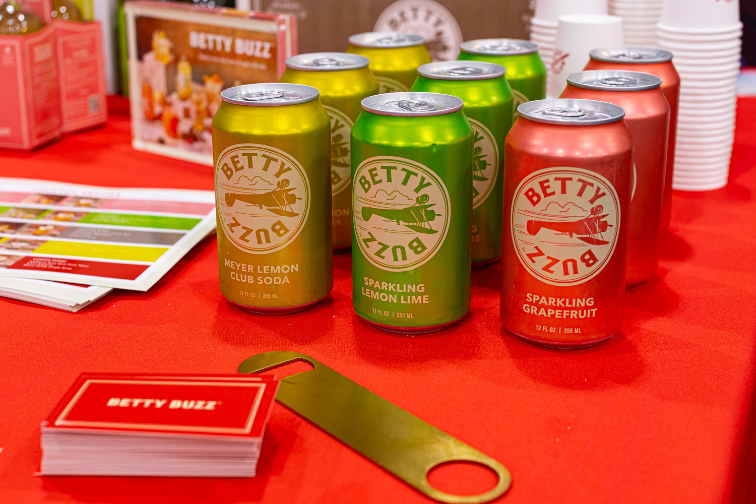 Cans of Betty Buzz sparkling lemon lime, grapefruit and water lemon soda sit on a red tablecloth with a bottle opener and cards