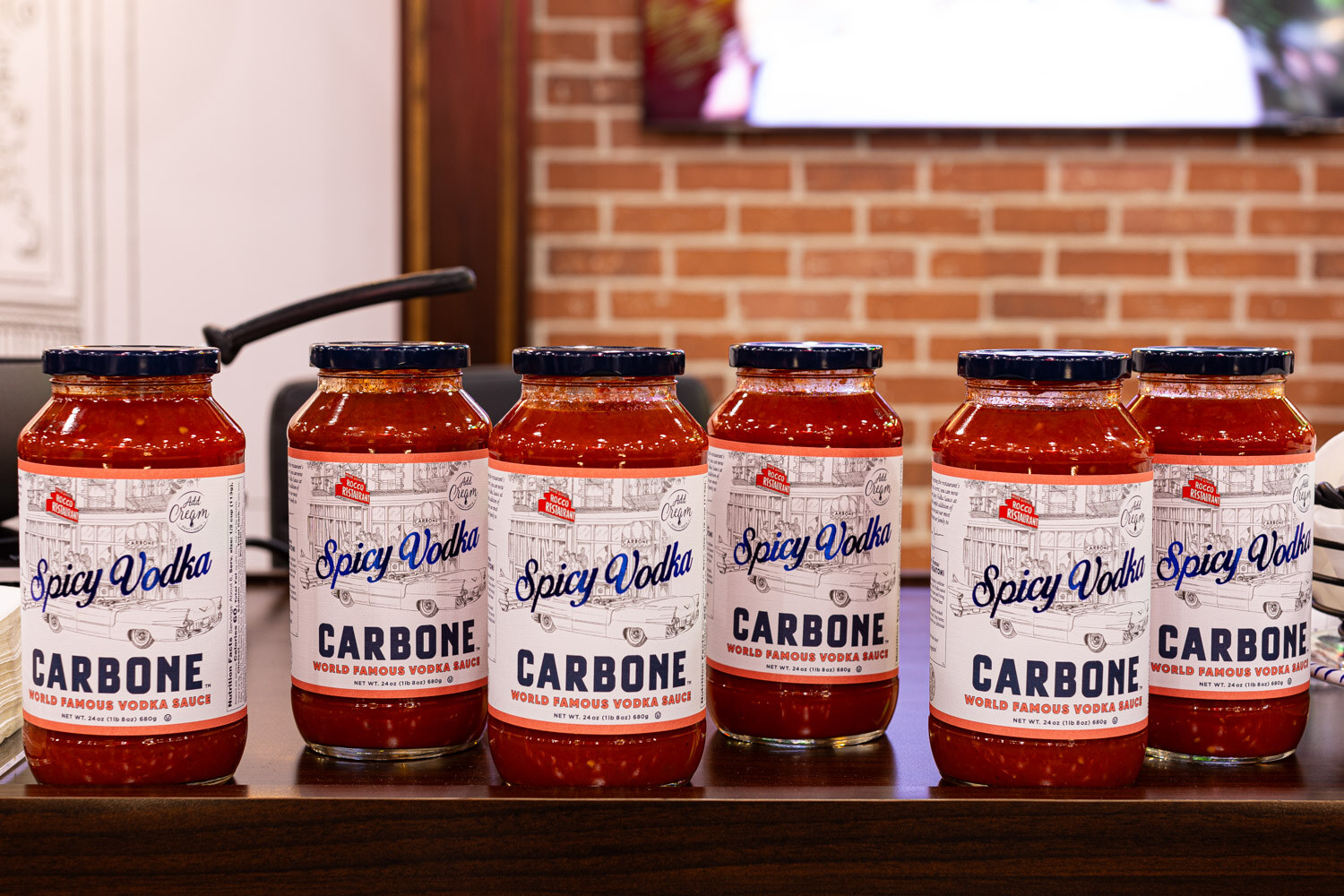 A row of Carbone Spicy Vodka sauce jars lines a wooden shelf in front of a brick wall
